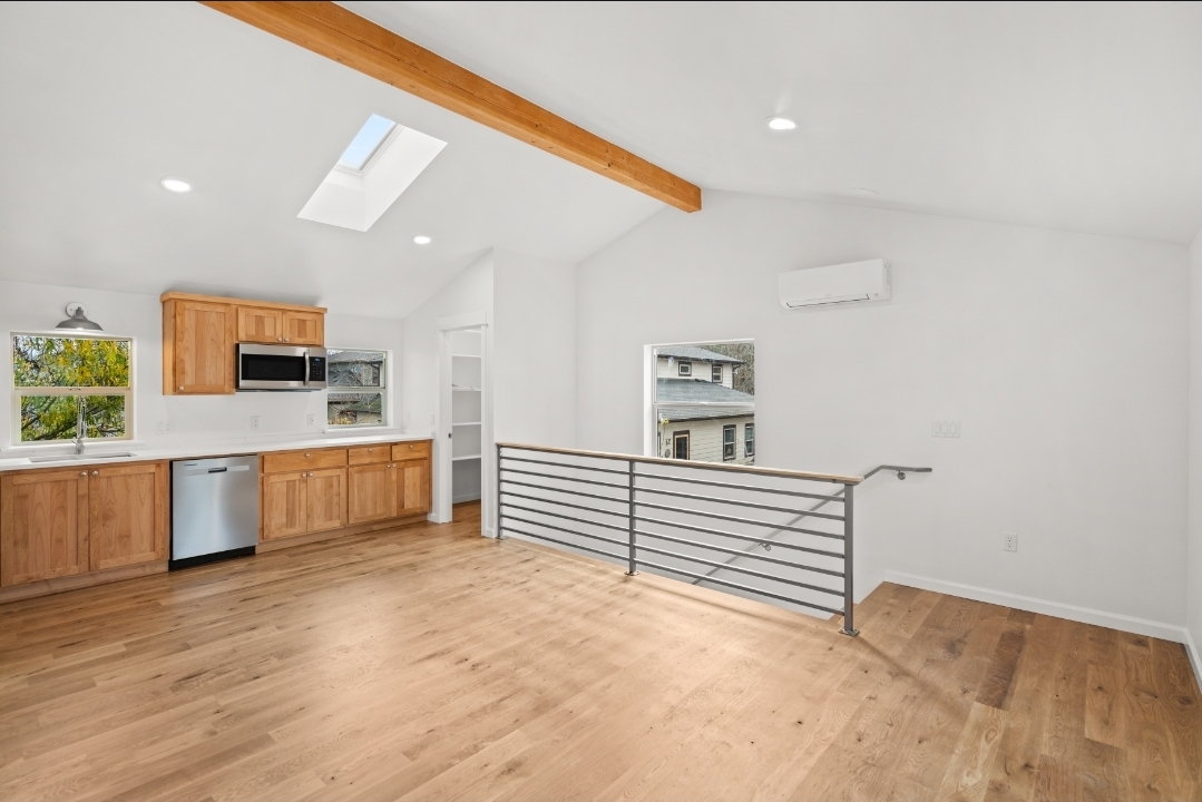 Open kitchen with wooden cabinets, white walls, skylight, and hardwood flooring, with a railing and small window on the right.