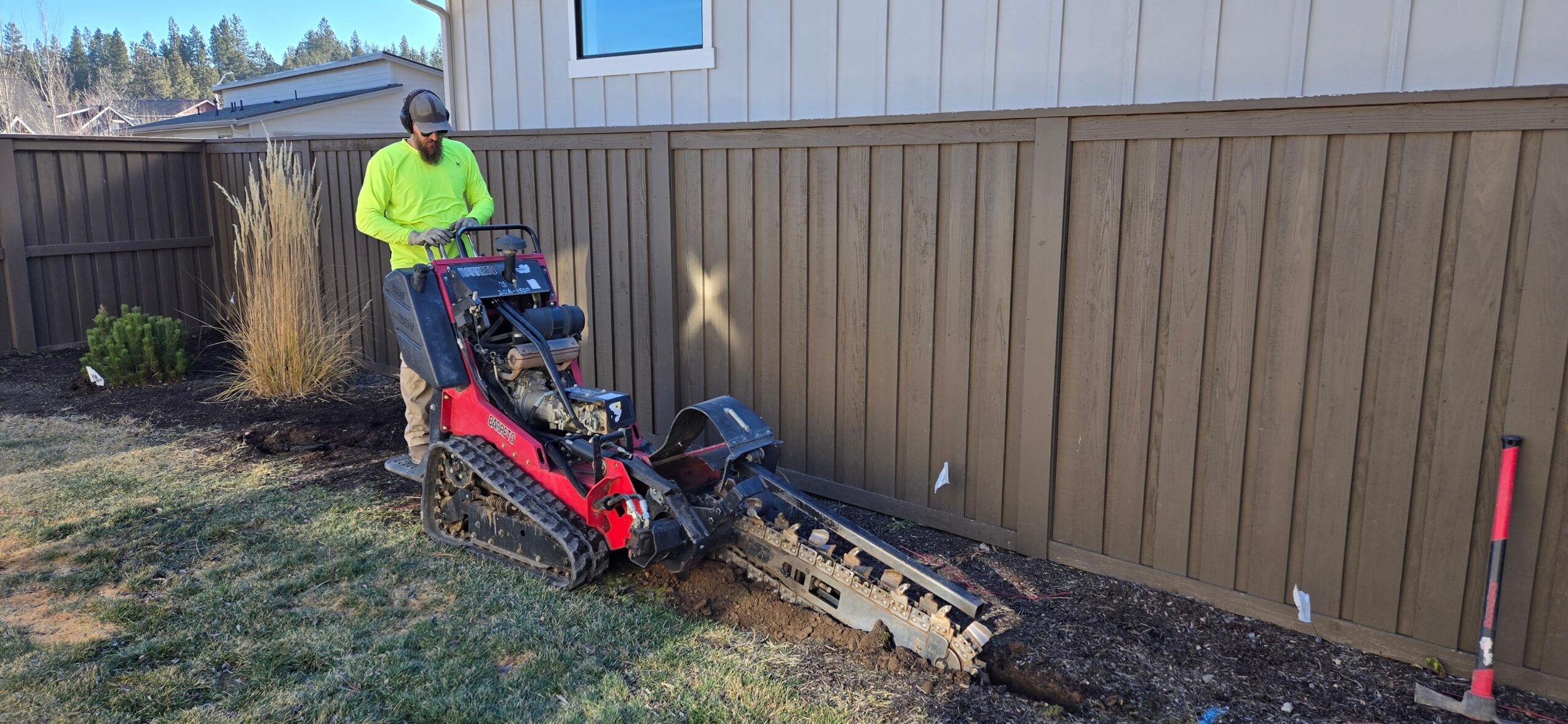 Person operating a small tracked excavator digging near a wooden fence in a yard, with a house in the background.