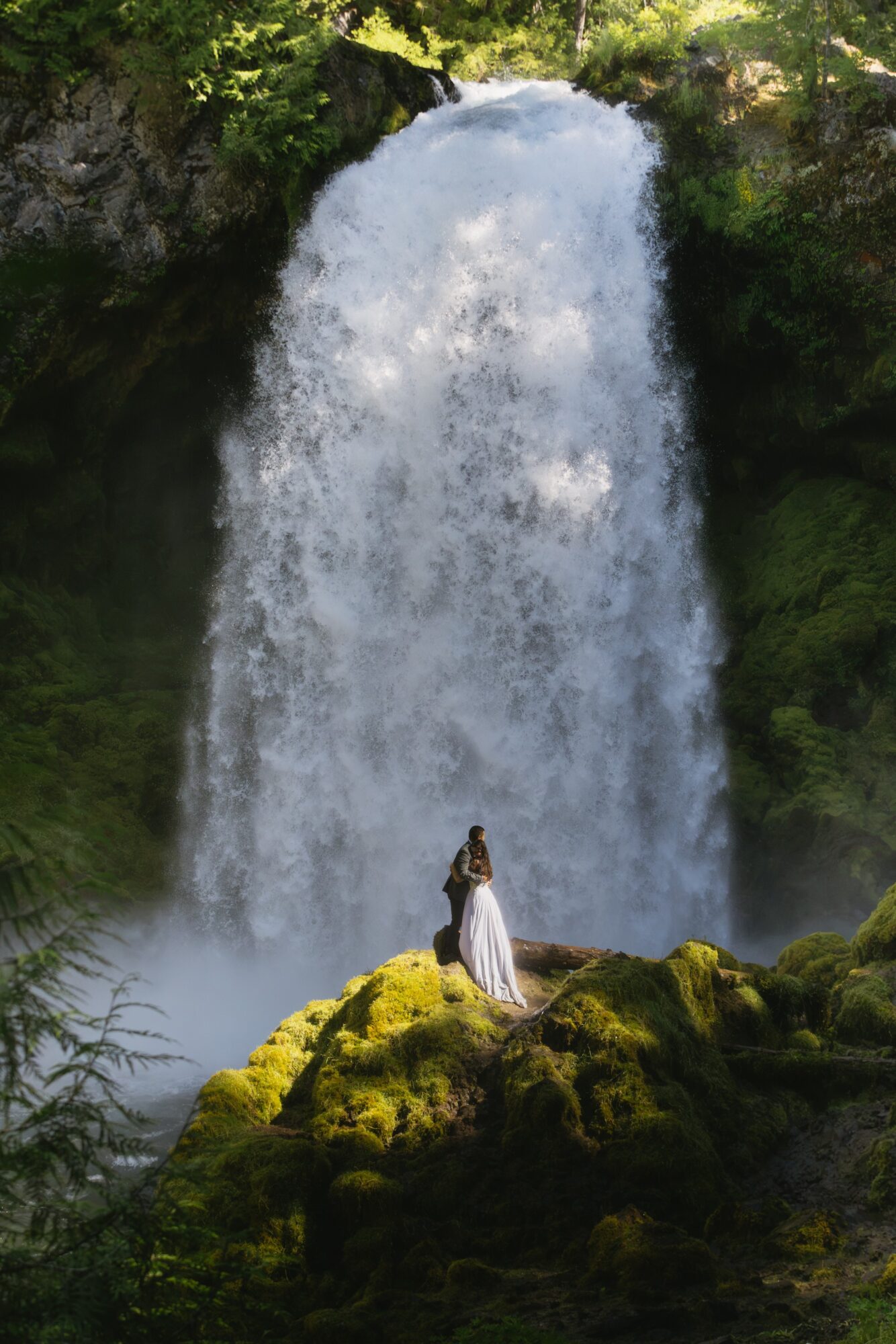 Person in a white dress standing on mossy rocks in front of a large waterfall surrounded by greenery.