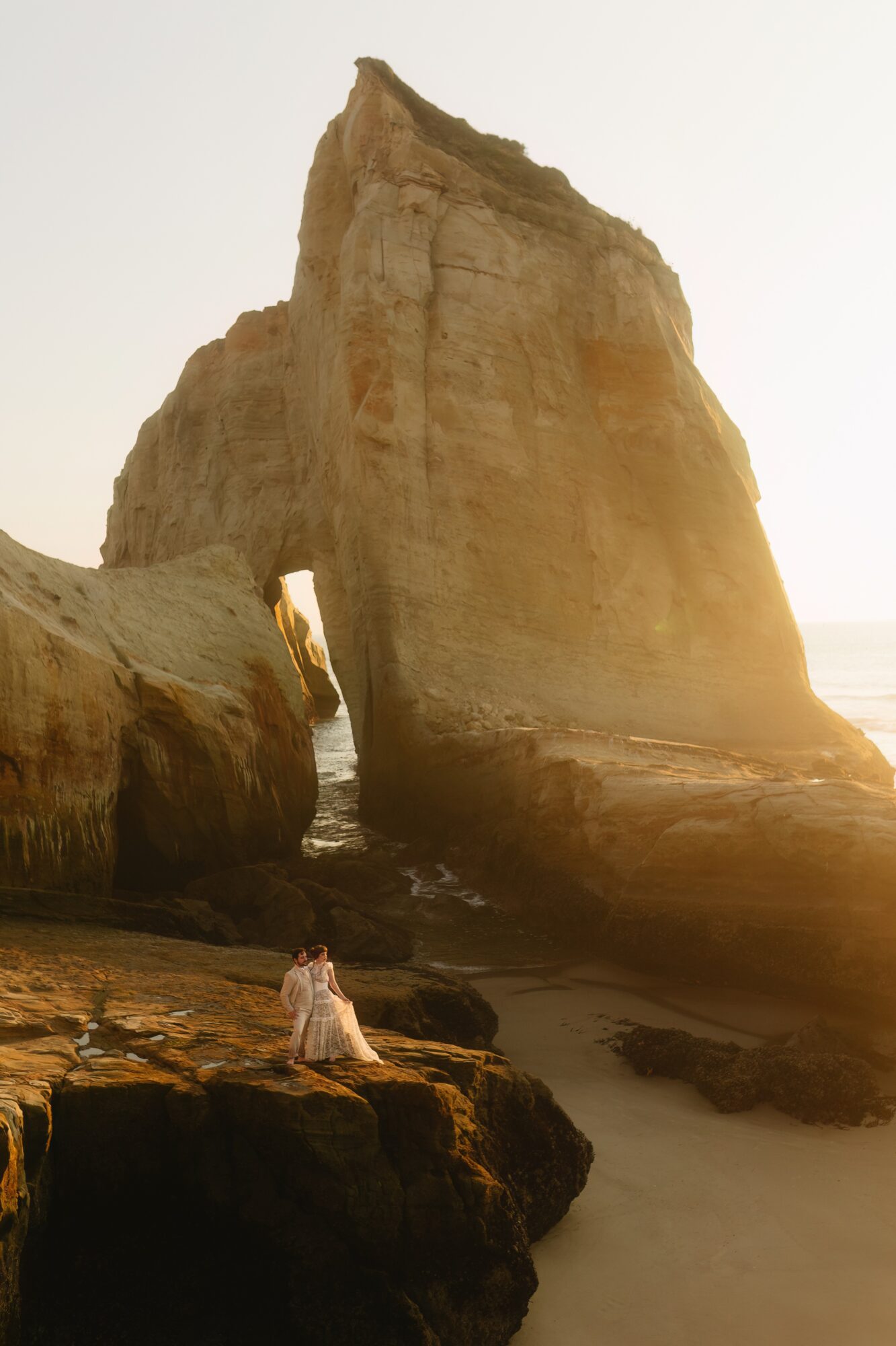Person sitting on rocks near large rock formation by the sea at sunset.