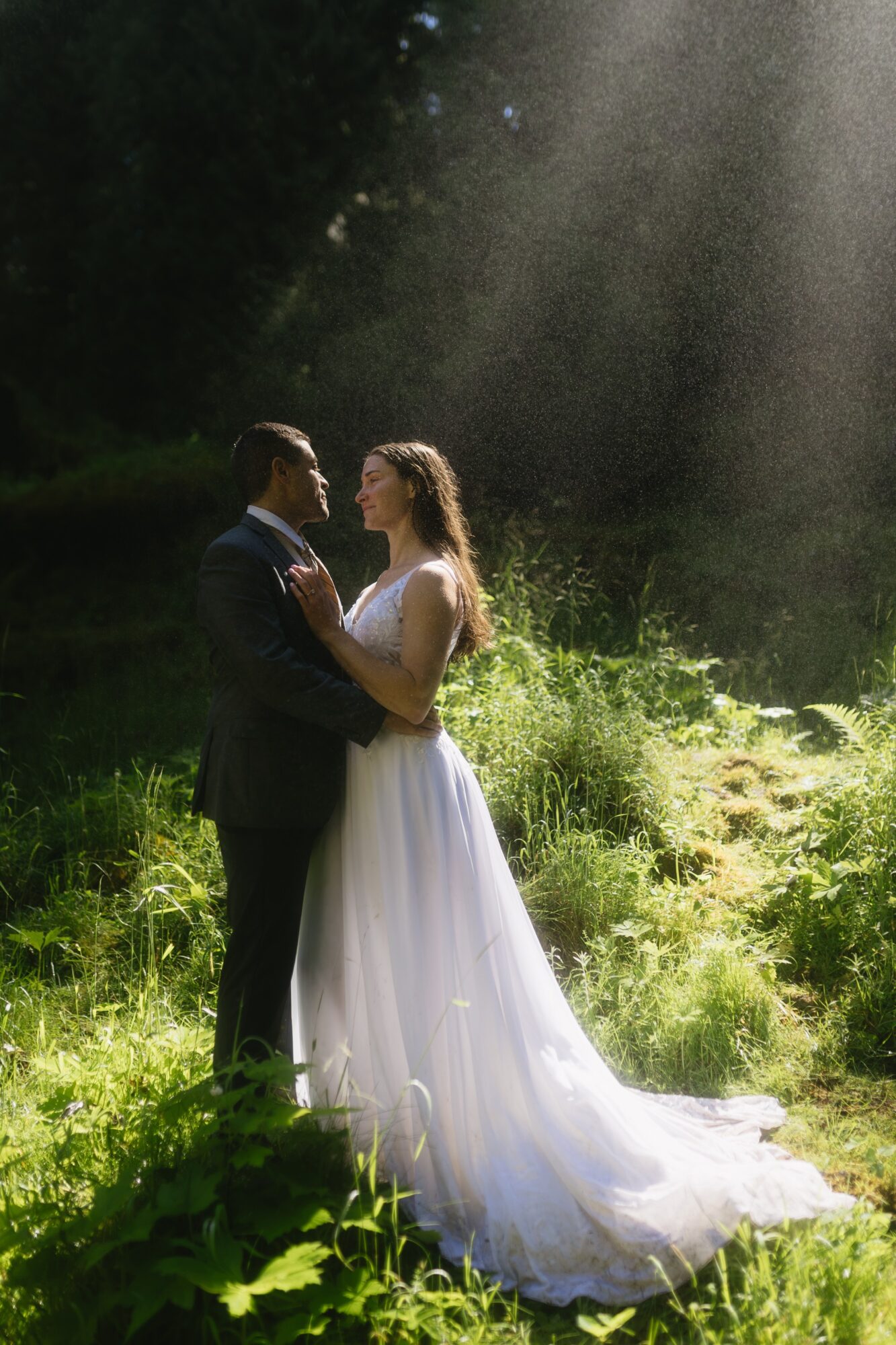 A couple in wedding attire standing in a sunlit outdoor setting with greenery, facing each other and holding hands.