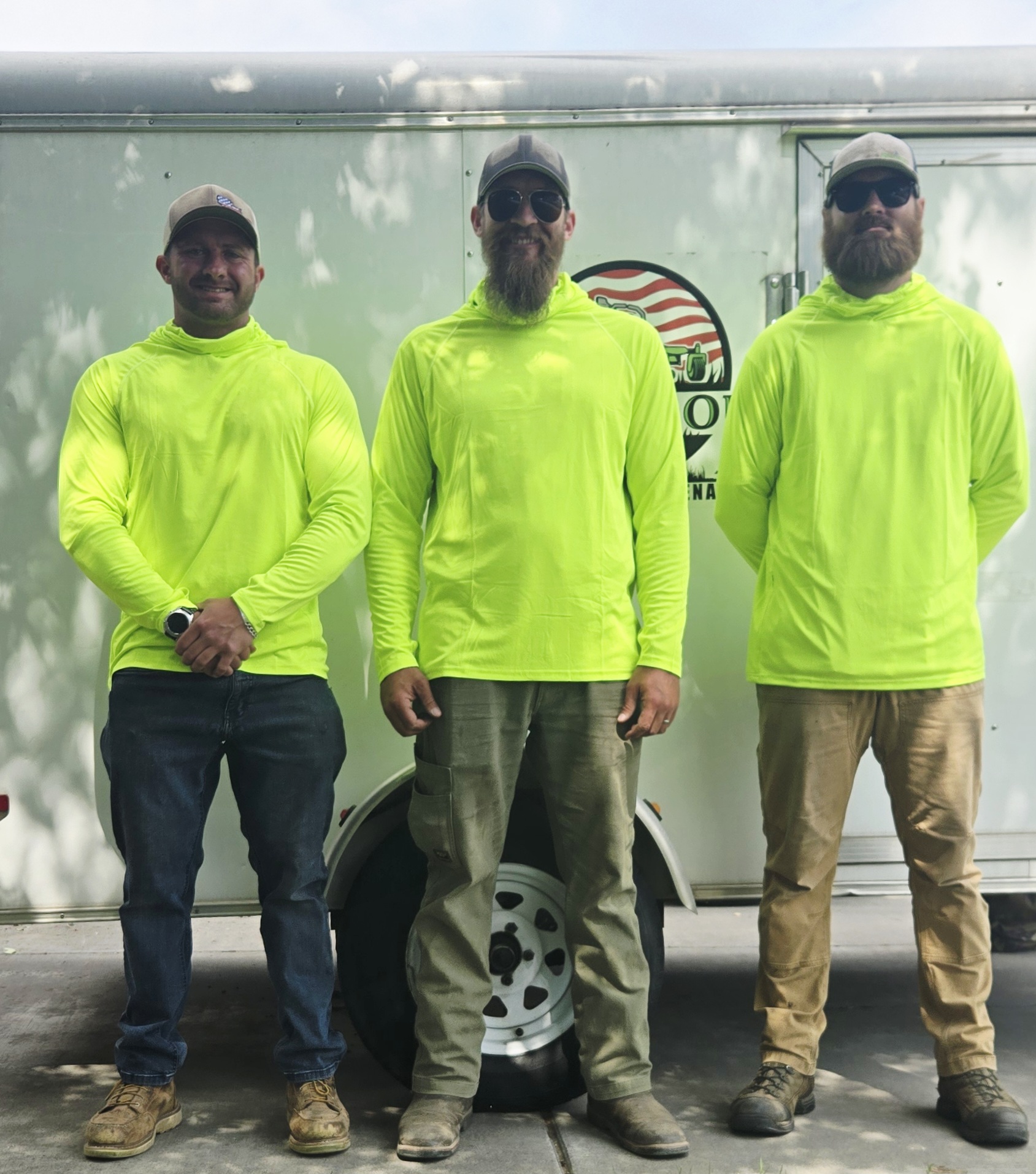 Three men in bright yellow jackets standing outdoors in front of a trailer with a logo, smiling, with trees and sky in background.