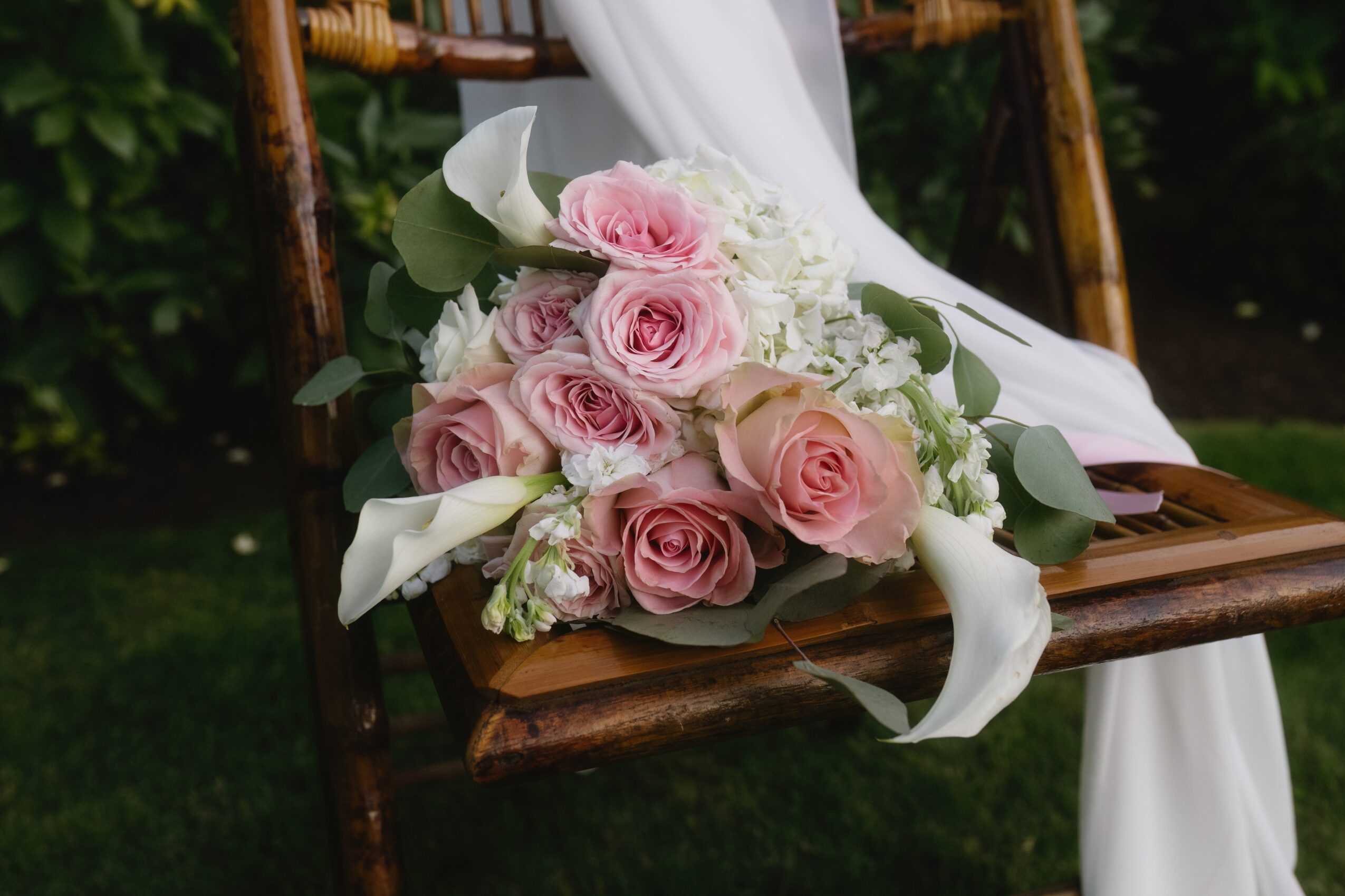Bouquet of pink and white roses with greenery and white ribbons on a wooden chair outdoors.
