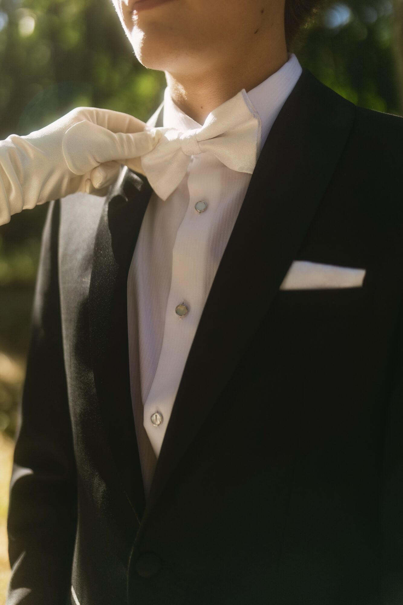 Person in formal tuxedo with white bow tie, outdoors with greenery background.