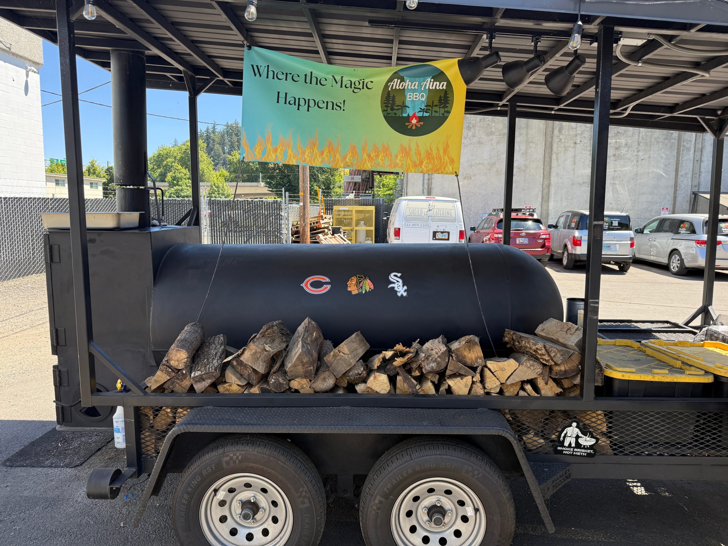 Wood logs stored in a black metal container on a trailer, with a sign above reading 'Where the Magic Happens!' and a parking lot in background.