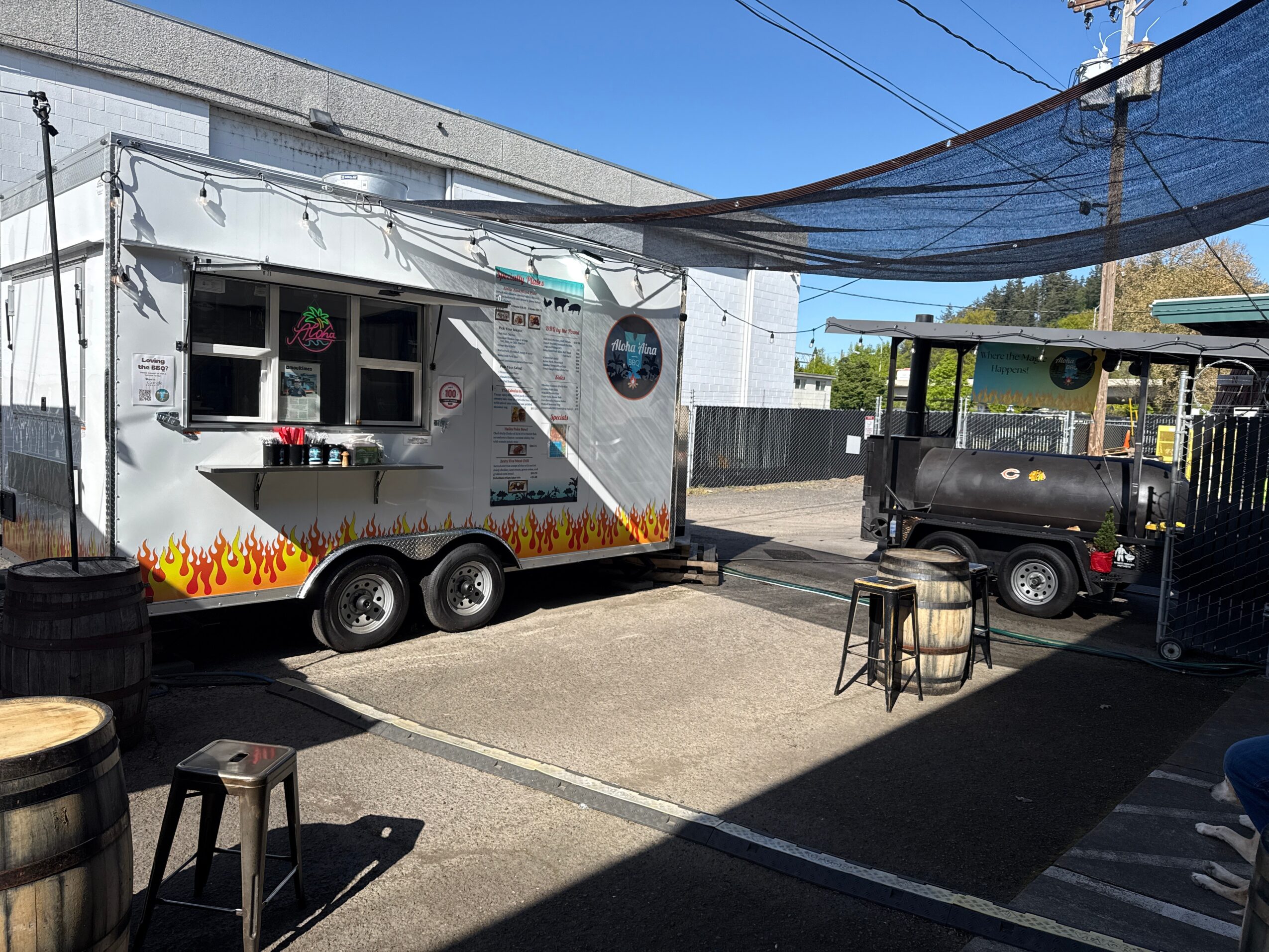 Food truck with flame design parked outdoors under a shade cloth, with stools and a small table nearby, on a sunny day.