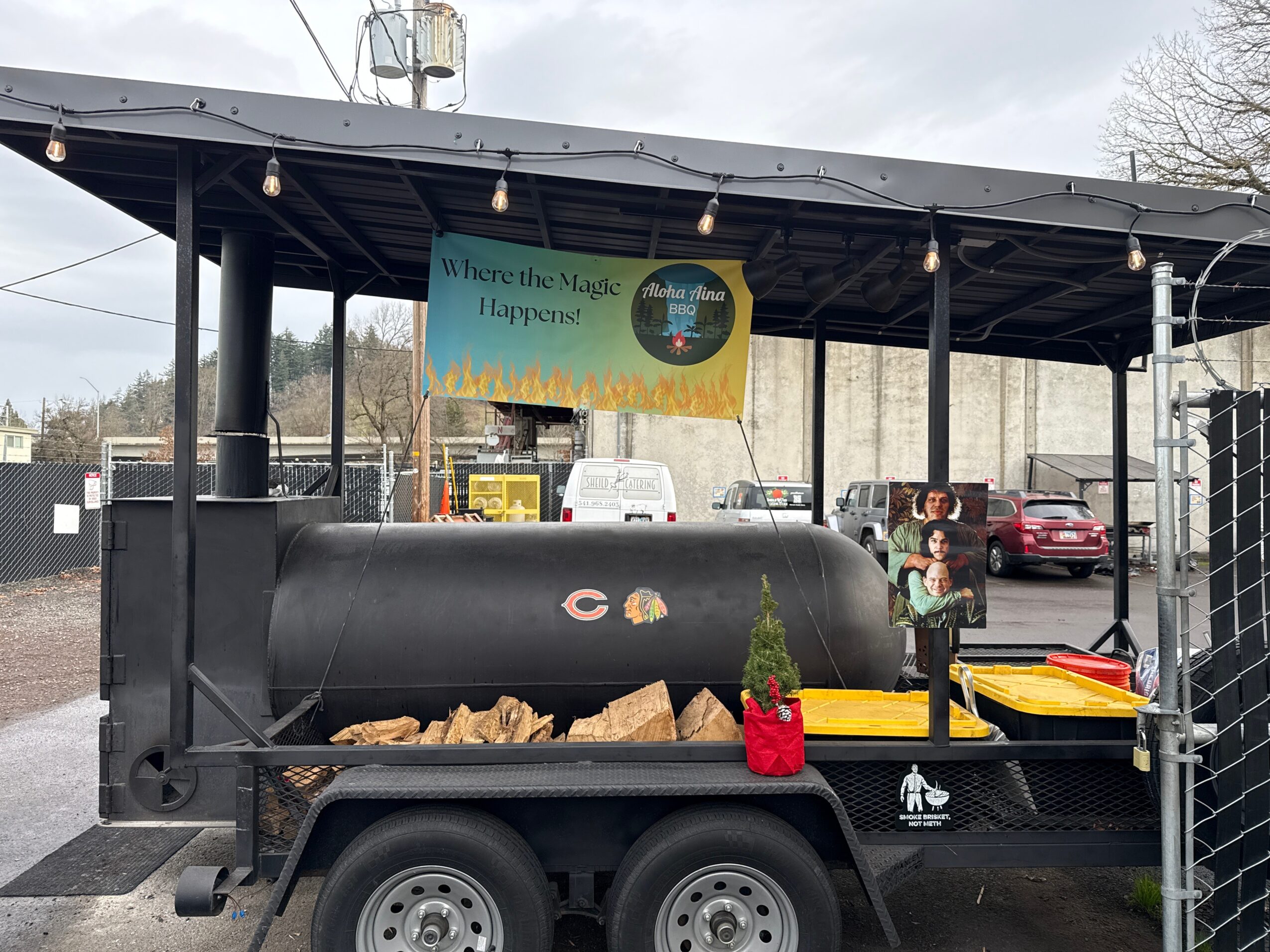 A food truck with a large black grill, wood logs, and a person standing nearby in a parking lot.