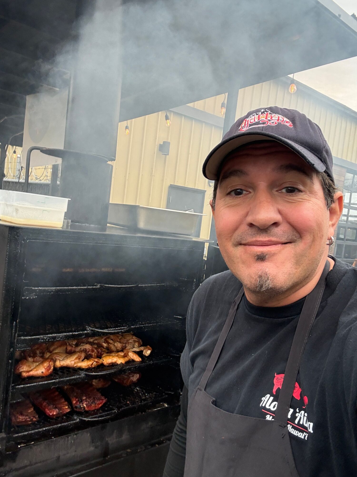 Man wearing a cap and black jacket grilling meat on barbecue with smoke rising, outdoors.