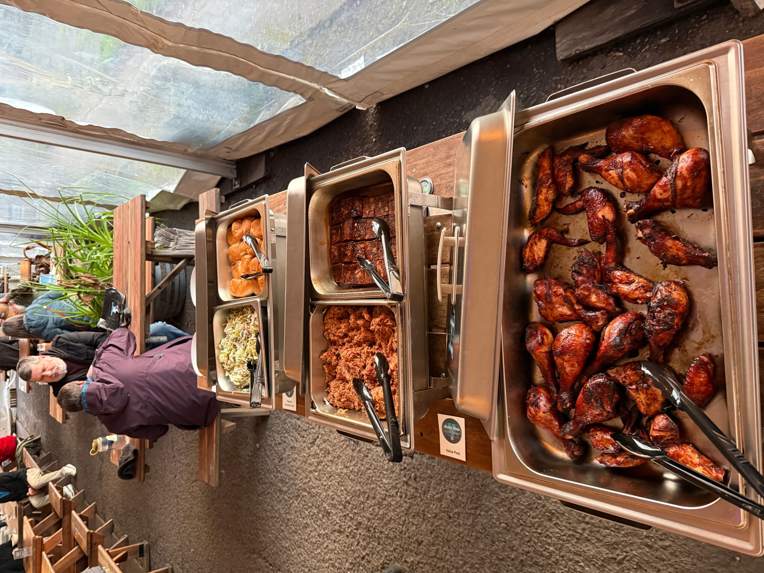 Buffet table with trays of grilled chicken, ribs, and side dishes, with people serving themselves outdoors under a canopy.