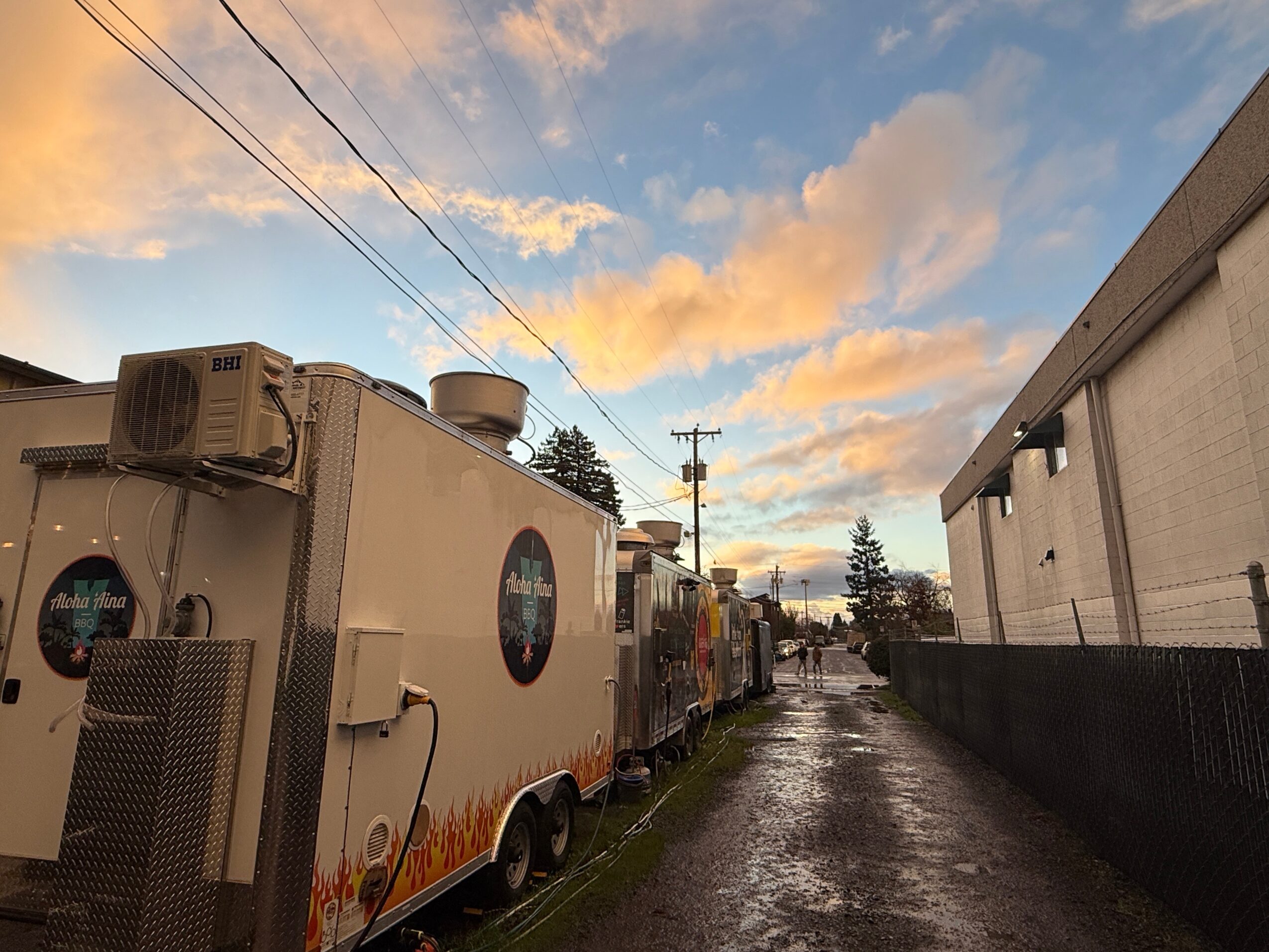A trailer with a circular logo parked on a wet street during sunset, with a building on the right and power lines overhead.