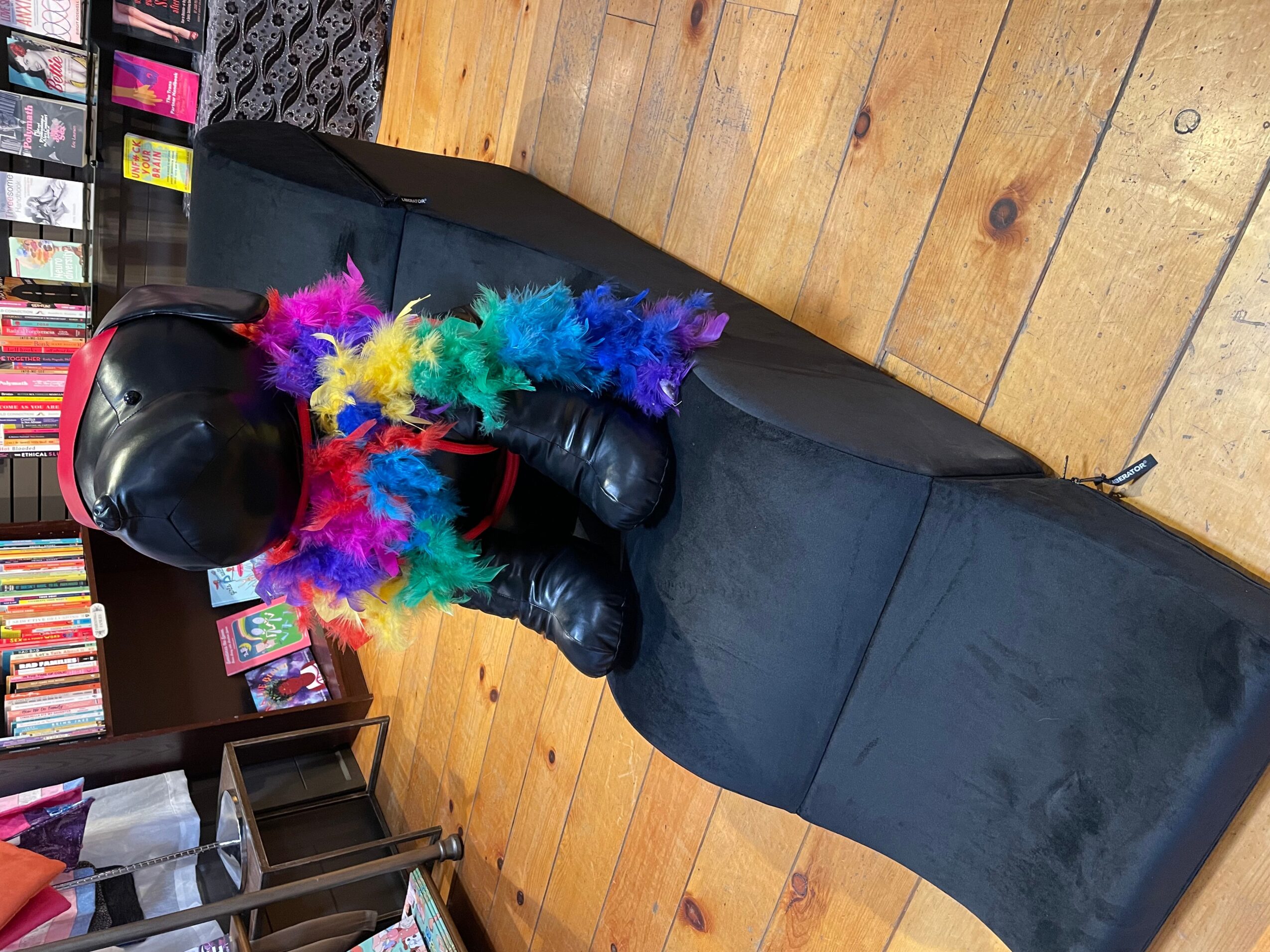Person in black leather outfit and helmet sitting on a black couch with colorful feather boa, wooden floor background.