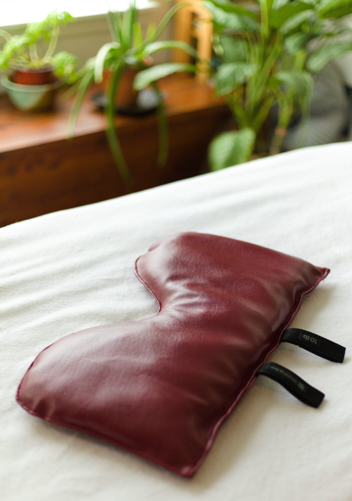 A maroon-colored sleep mask with black elastic straps rests on a white bed sheet, with green plants in the background.