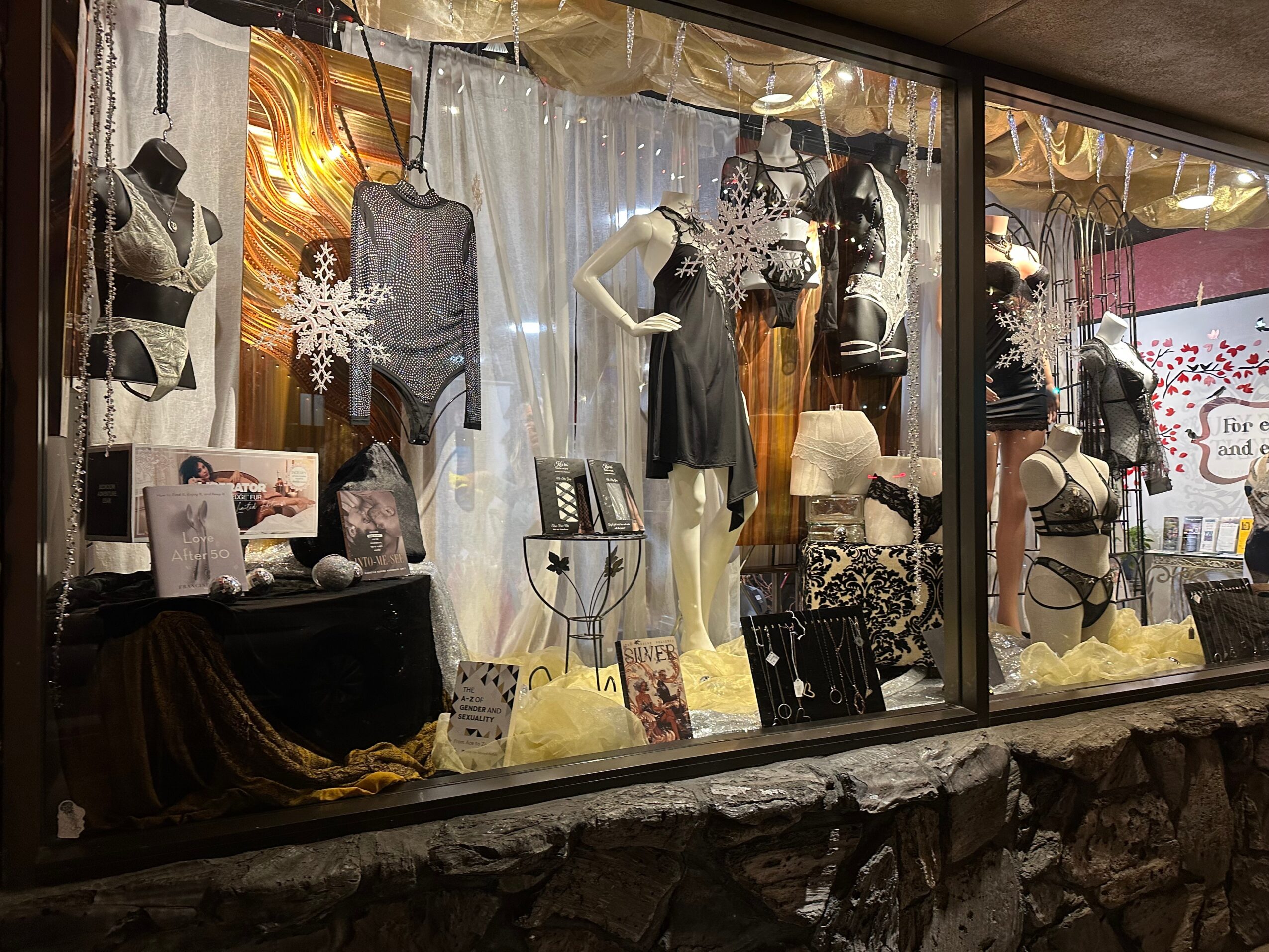Store window display with mannequins dressed in lingerie, holiday decorations, and a snowy theme, with curtains and decorative snowflakes.