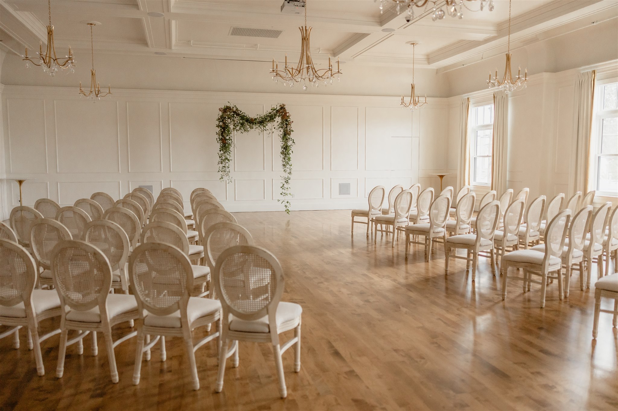 Empty wedding ceremony room with rows of white chairs, a floral arch, and large windows, bright and elegant.