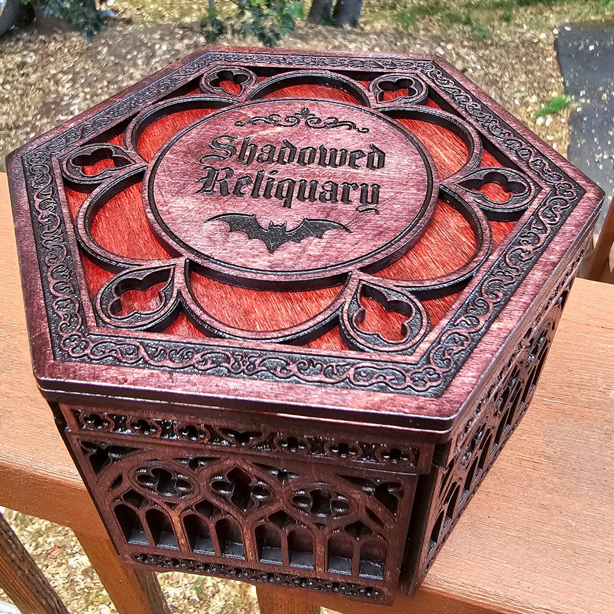 Decorative wooden box with intricate carvings and a central label reading 'Shadowed Religiousry' with a bat symbol, outdoors.