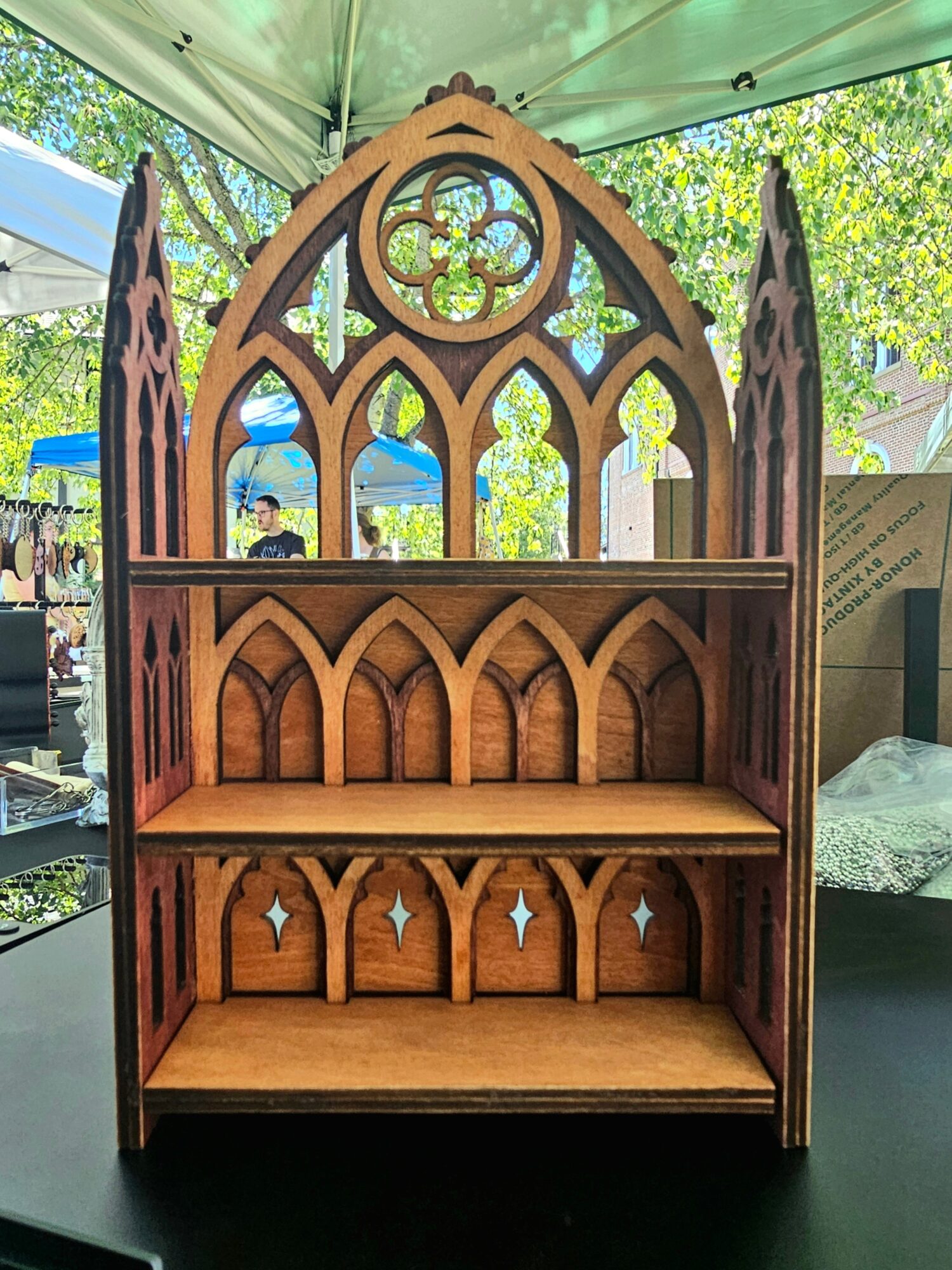 Wooden Gothic-style shelf with intricate arched designs, three shelves, set outdoors with trees and tents in background.