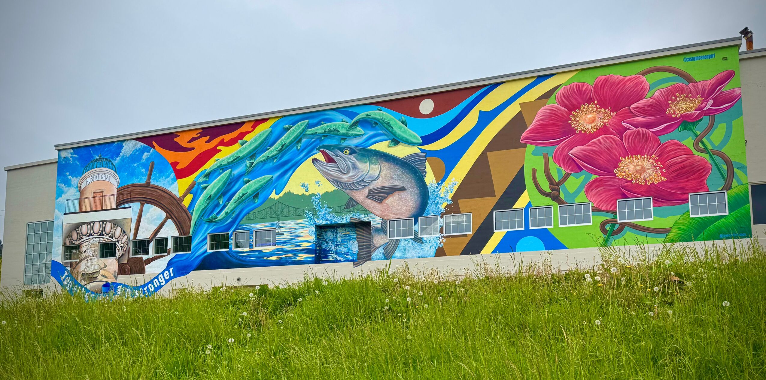 Colorful mural with a fish, flowers, and abstract shapes on a building wall, with grass in the foreground.