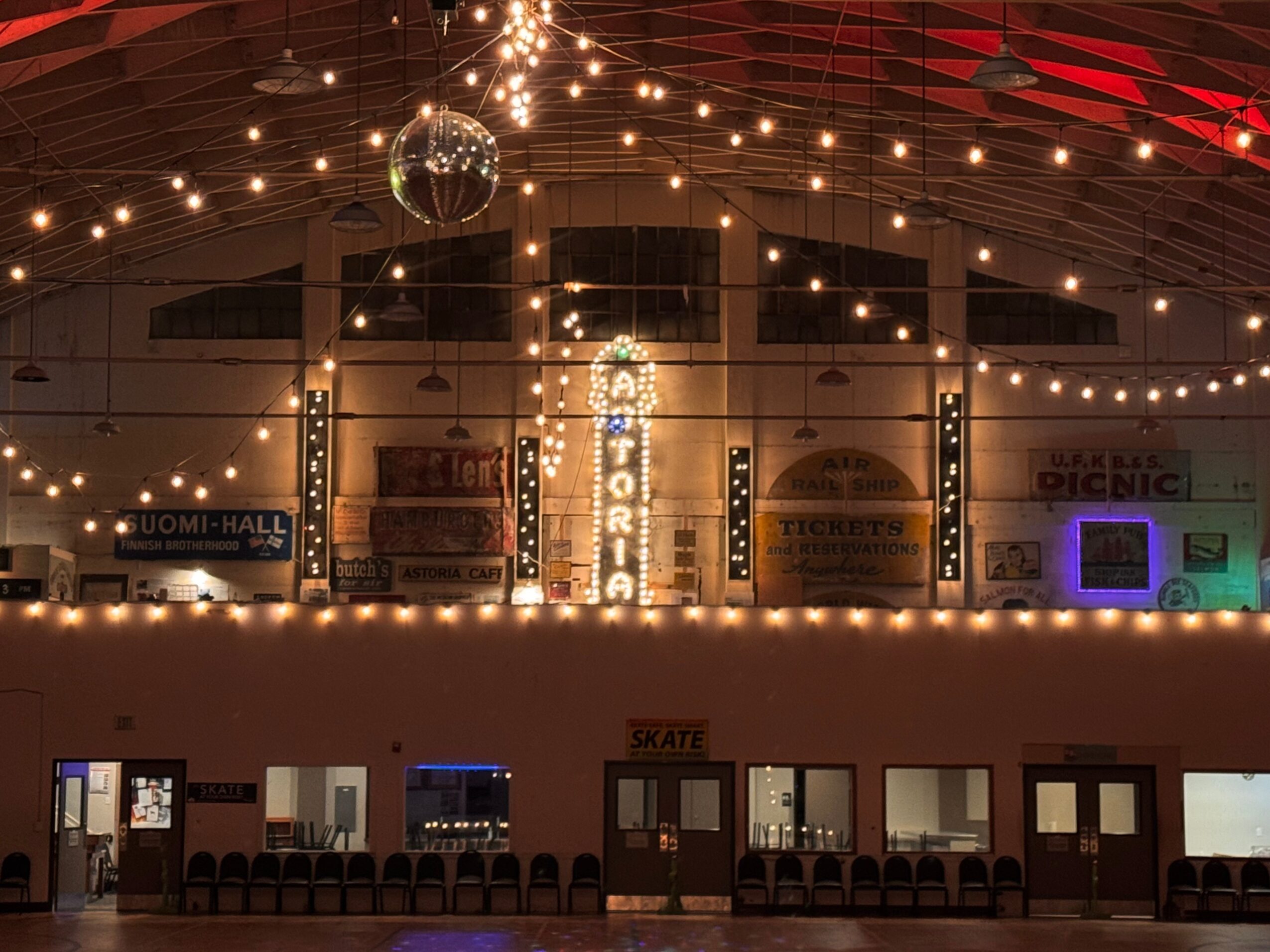 Indoor space decorated with string lights, a disco ball, and illuminated signs, with seating and a stage area.