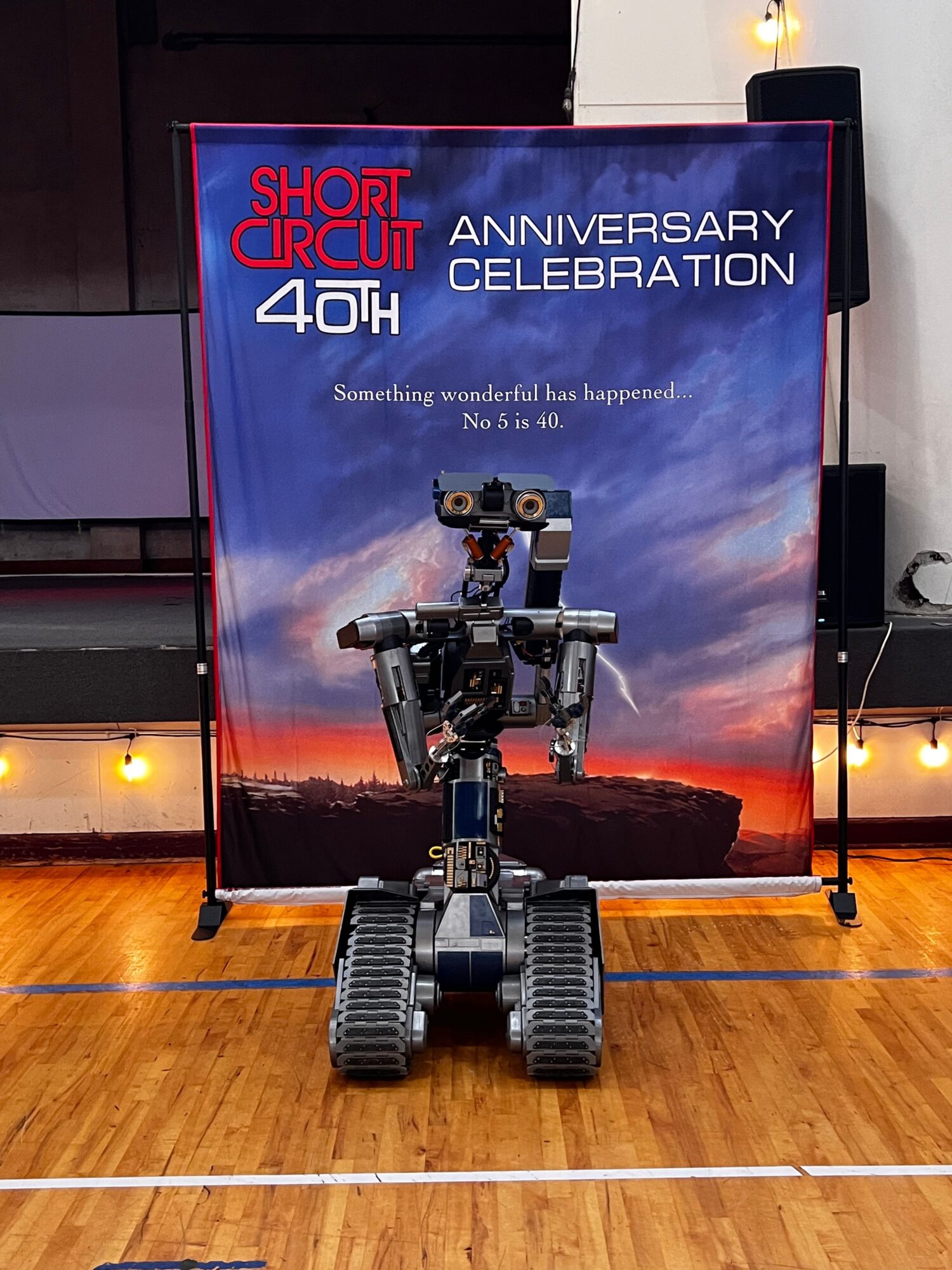 Robotic arm with wheels on a wooden floor in front of a colorful banner celebrating the 40th anniversary of Short Circuit. Alt text word count: 24