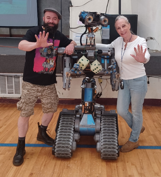 Two people, a man and a woman, stand on either side of a robot with tank tracks, waving and smiling.