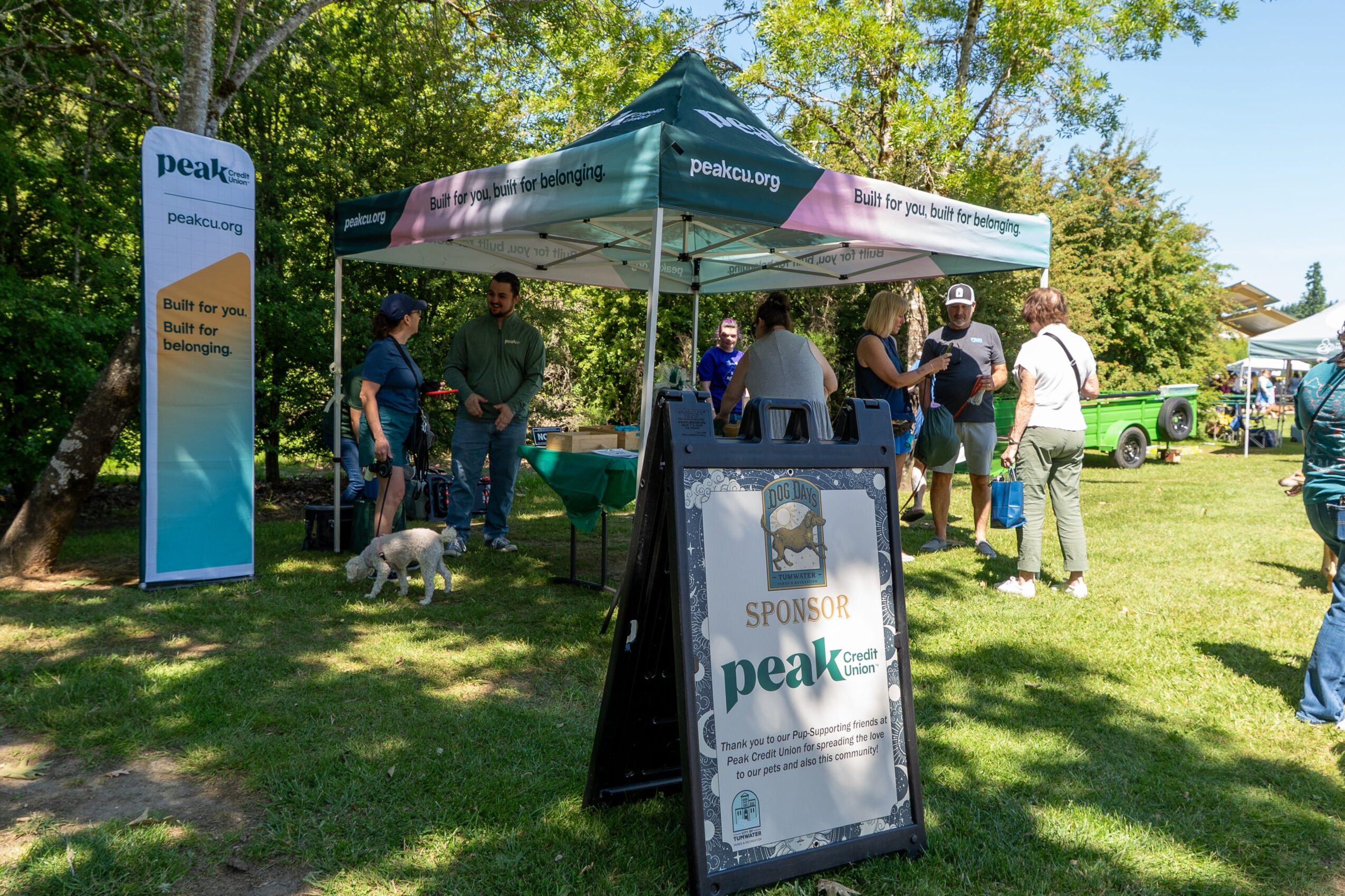 People gather around a tent at an outdoor event with trees and a grassy area, some holding leashes with dogs.