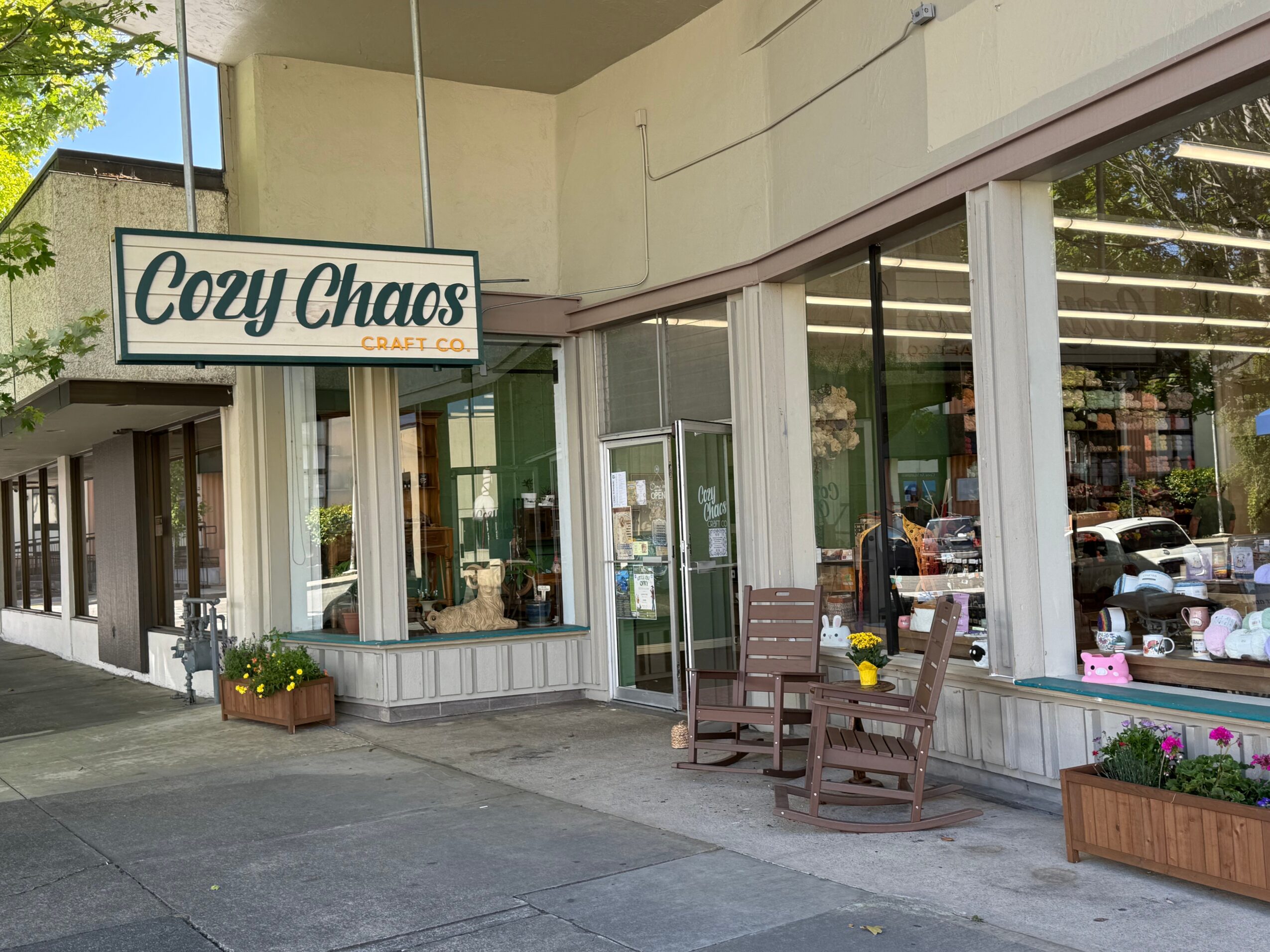 Storefront with a sign reading 'Cozy Chaos,' outdoor seating with chairs and flowers, large glass windows showing interior.