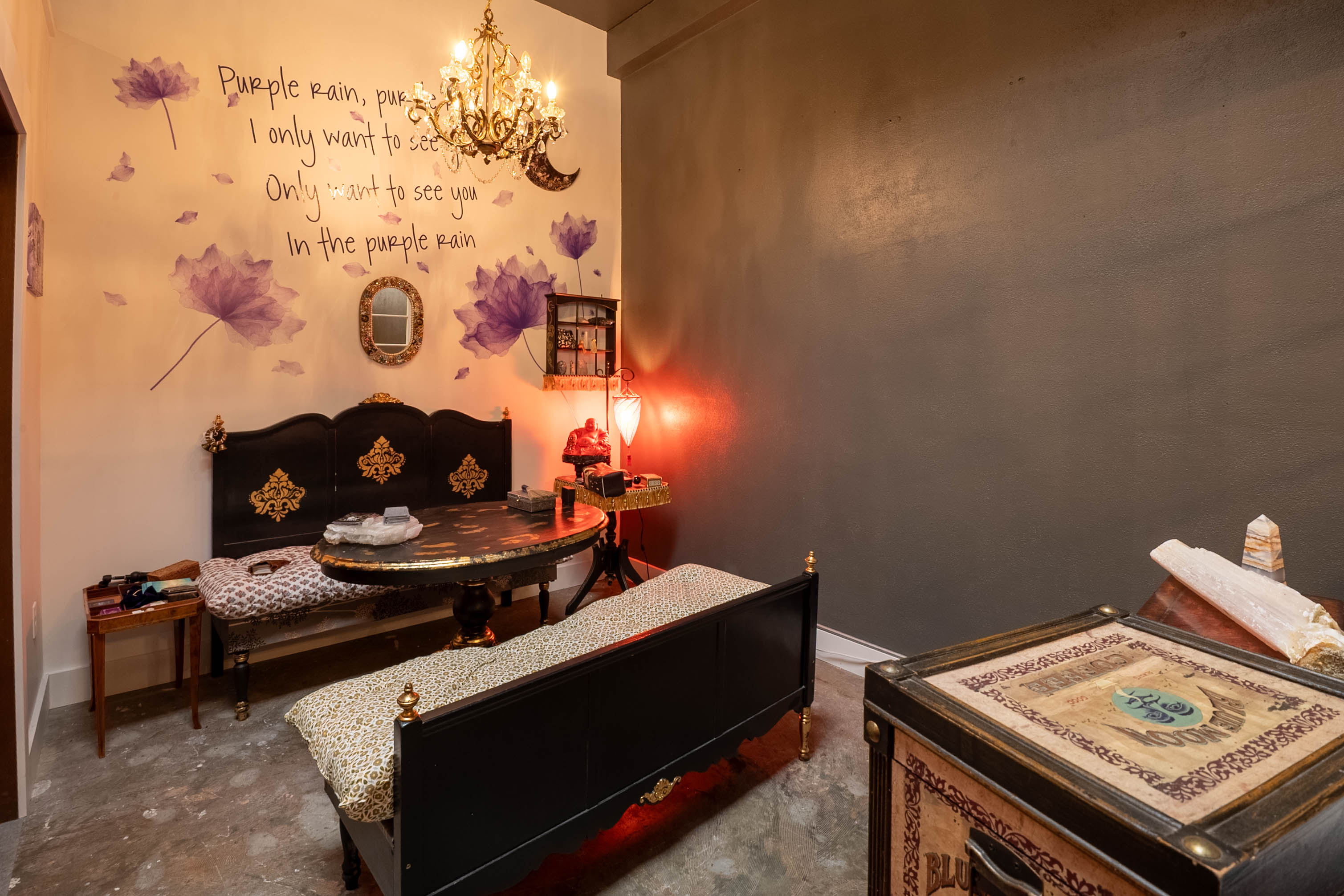 Room with a black sofa, a small table, a chandelier, and a wall with purple flower decorations and writing.
