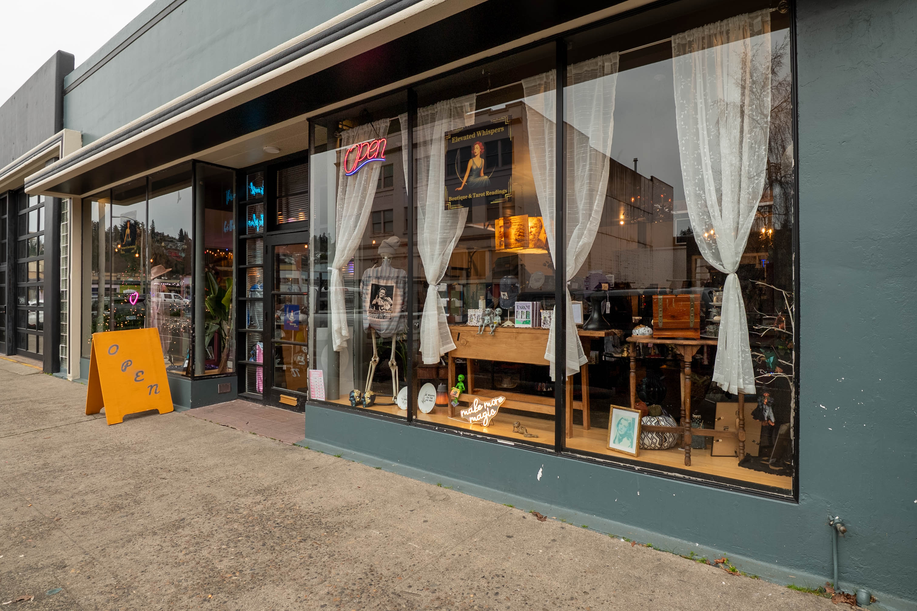 Storefront with large glass windows, white curtains, and a yellow sign outside, showing interior furniture and lighting fixtures.
