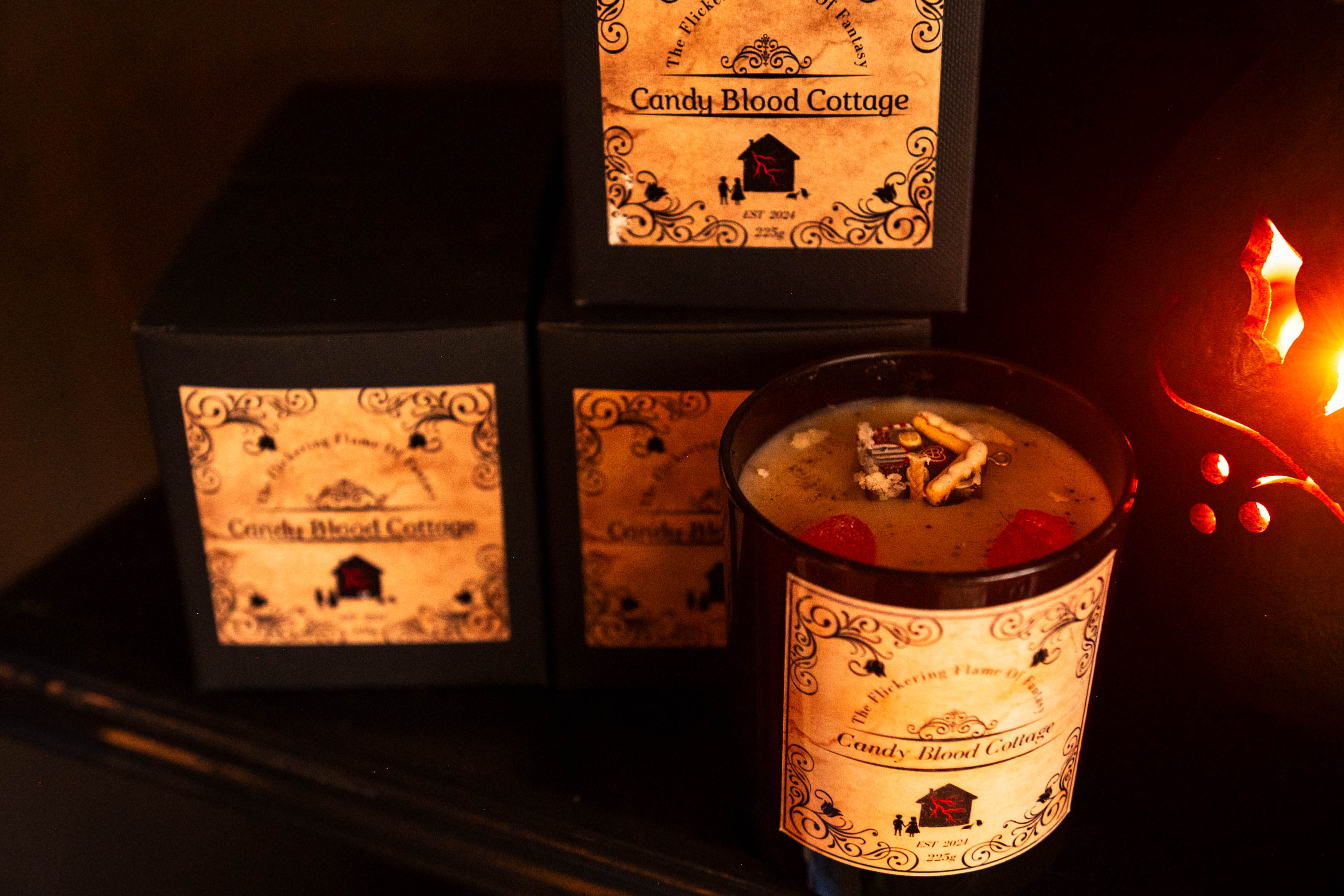 A candle in a glass holder with a label, surrounded by black boxes with similar labels, in a dimly lit setting.