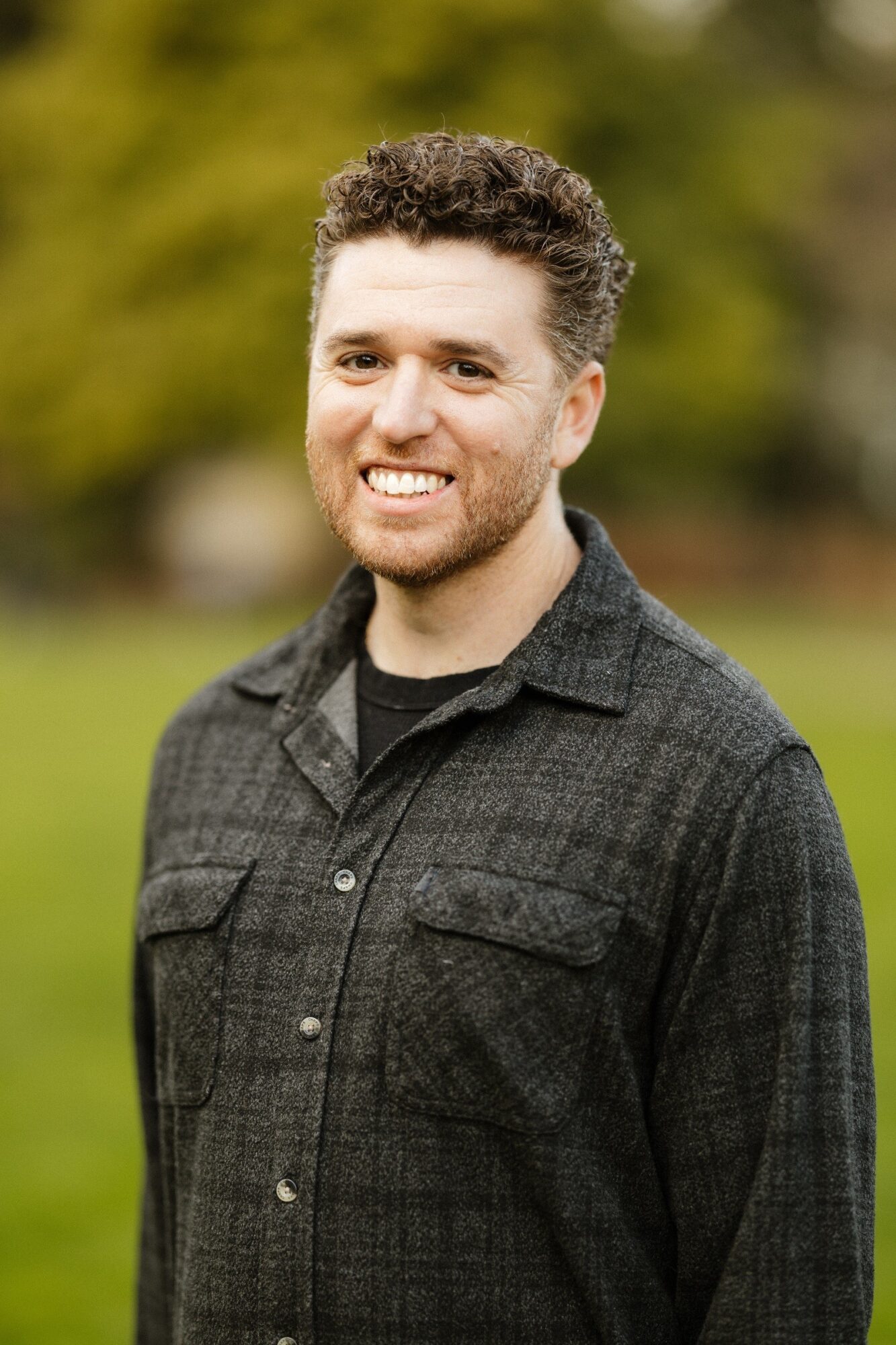 Smiling man with curly hair and beard outdoors, wearing a dark button-up shirt, blurred green background.
