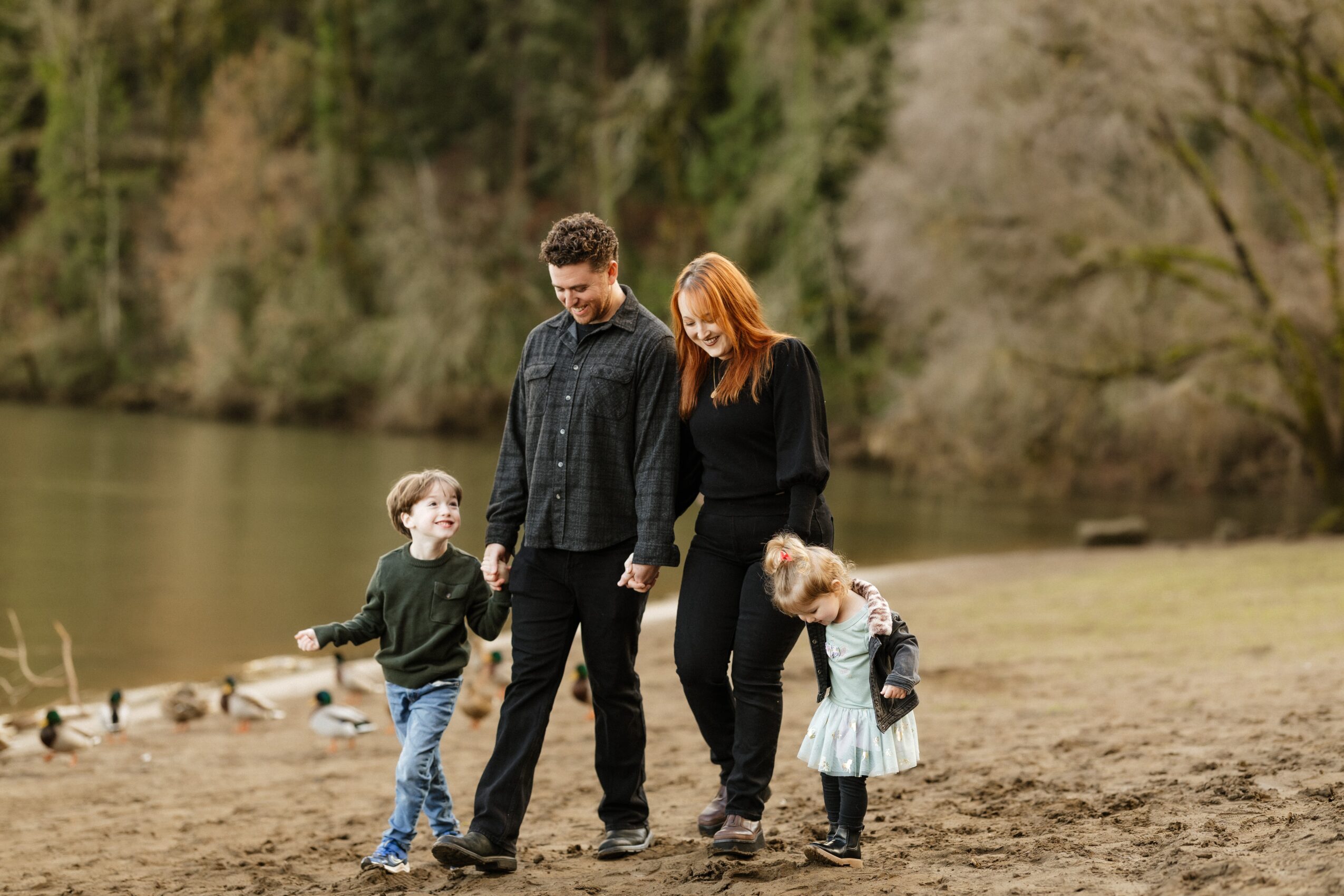 Family of four walking on a grassy area near a river with trees in the background, holding hands and enjoying outdoors.