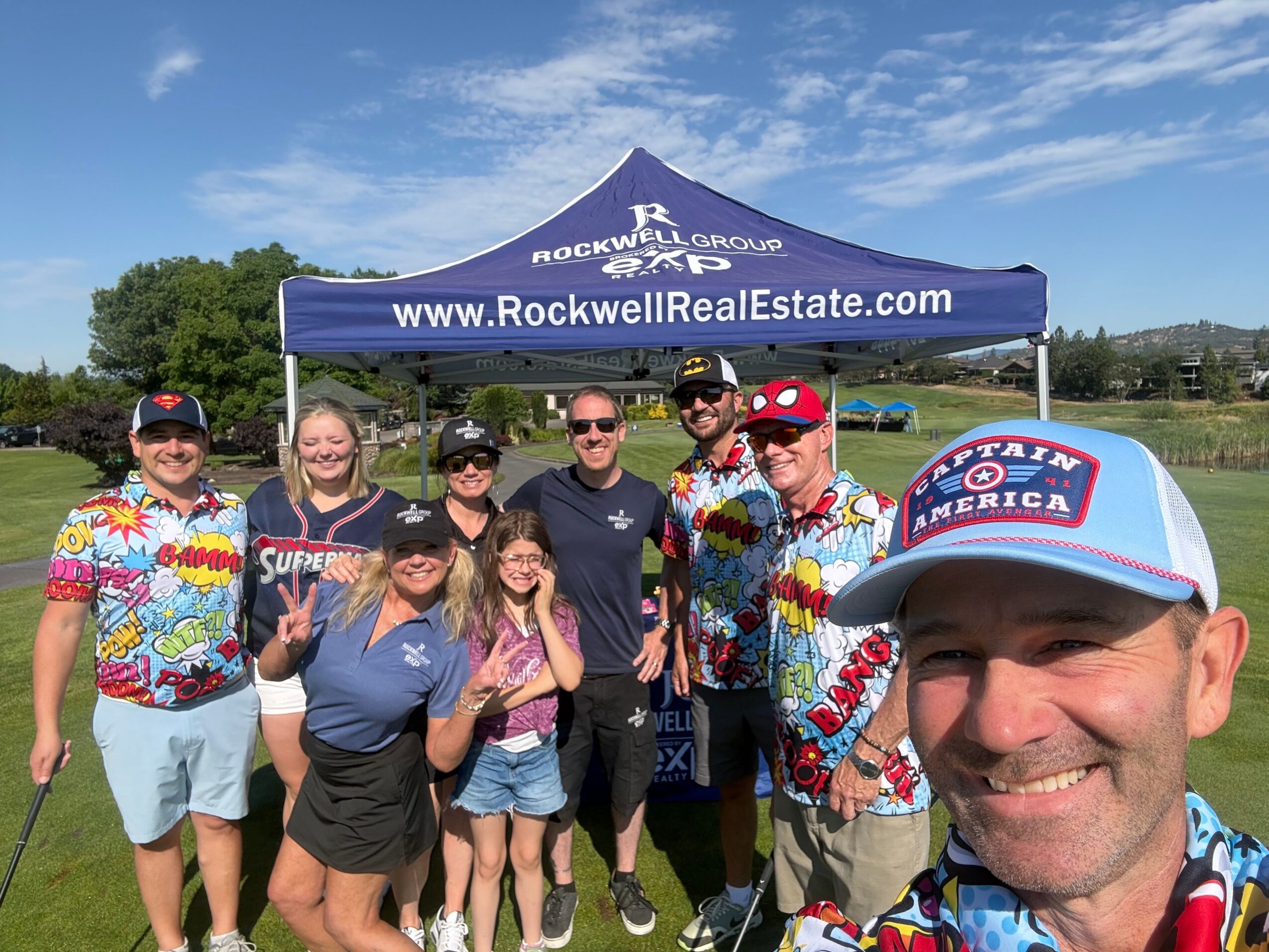 Group of people outdoors holding a banner, some wearing colorful shirts and sunglasses, with a blue sky background.