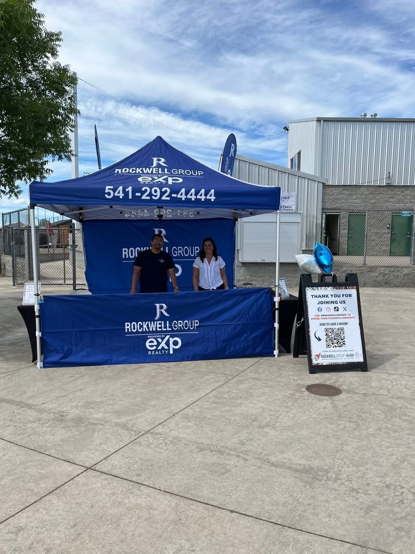 Two people stand behind a blue Rockwell Group booth with a canopy and table, outdoors on a sunny day.