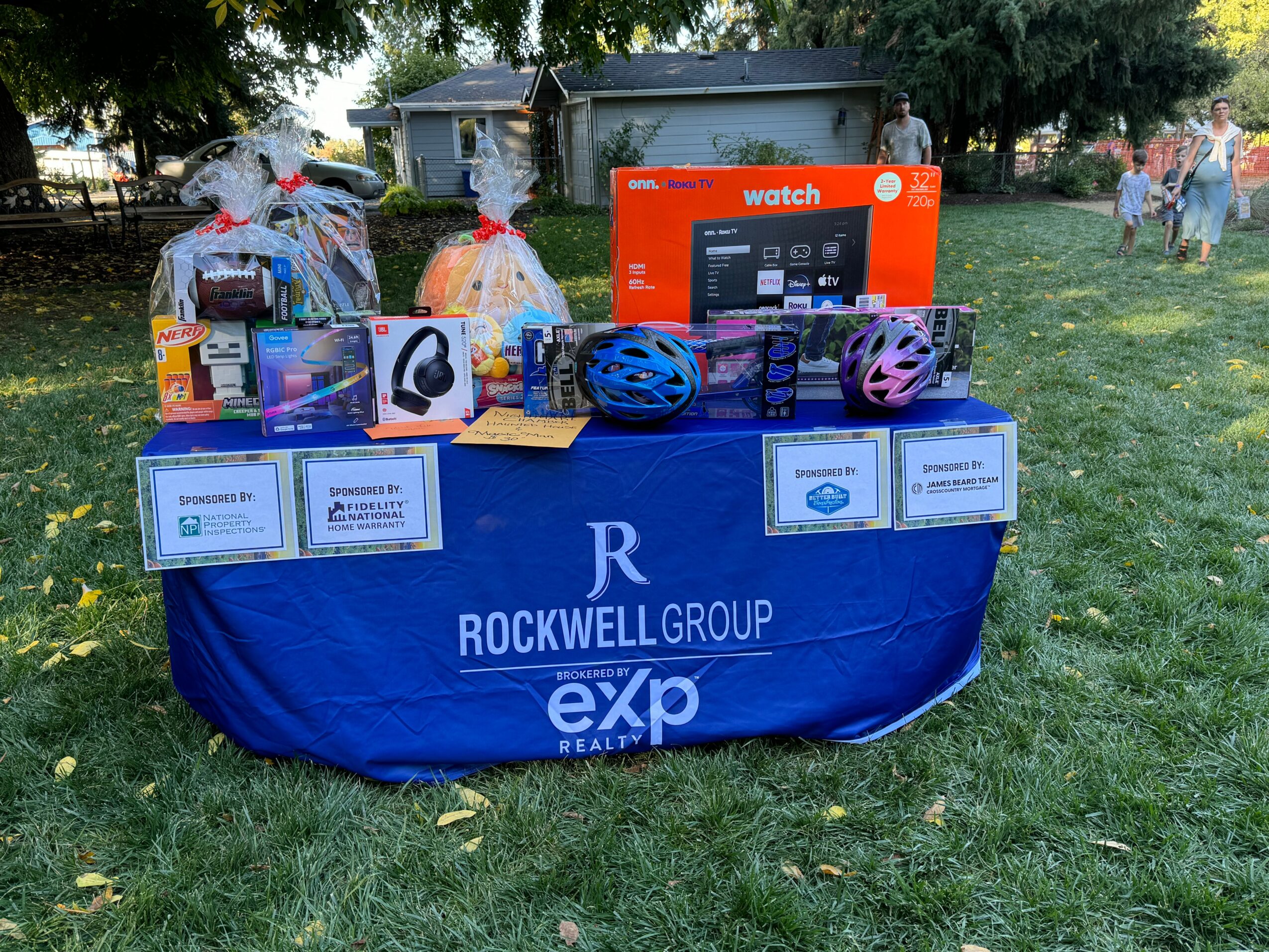 Table with prizes including headphones, helmets, and electronics, set outdoors on grass with trees and people in background.