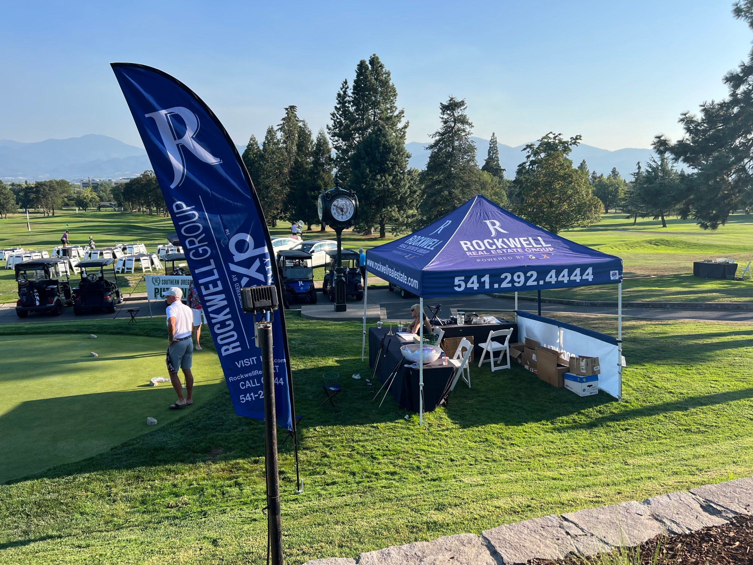Outdoor event setup with a blue tent and flag on a grassy area, trees, and mountains in the background.
