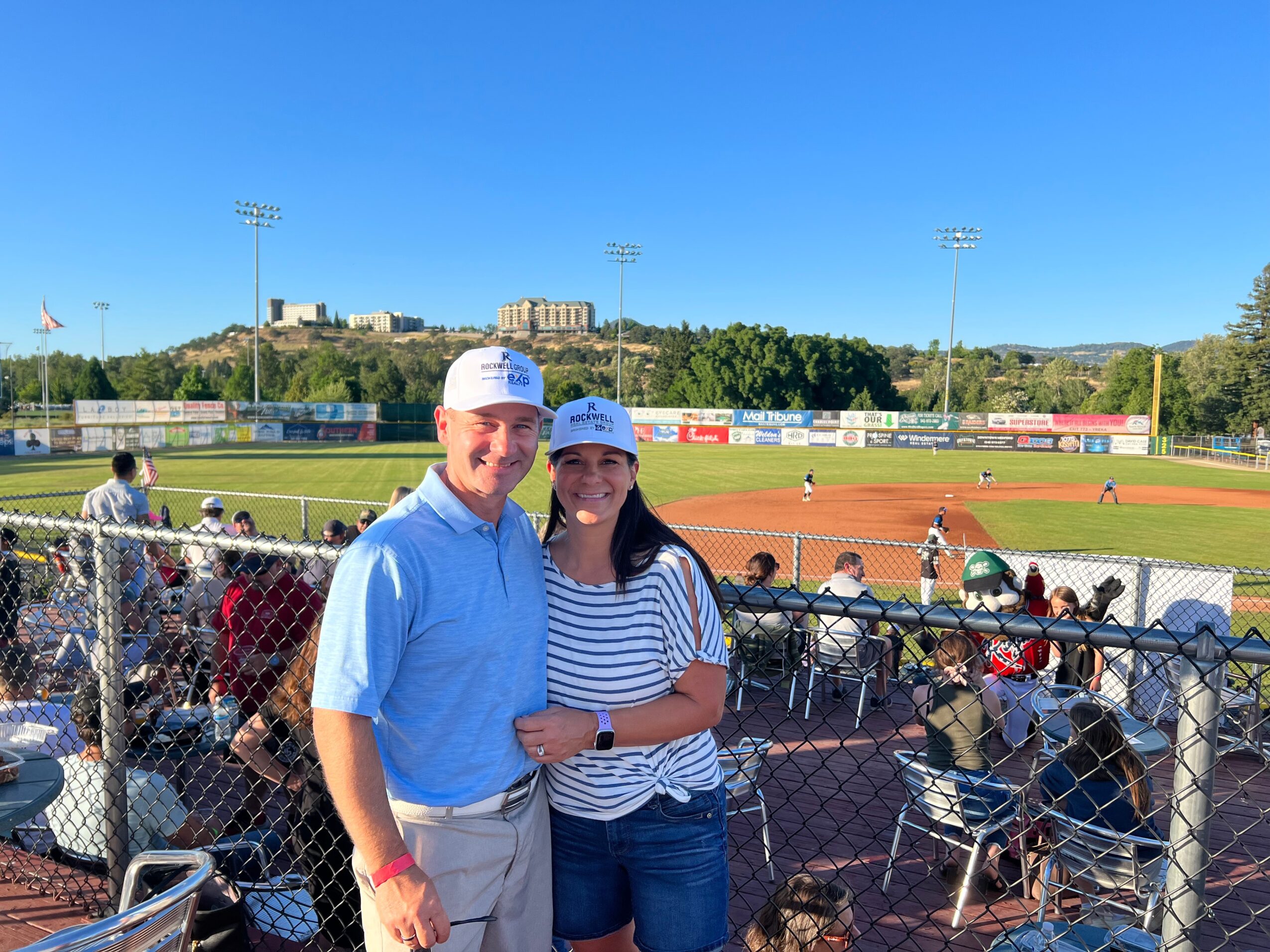 Two people smiling at a baseball game, standing behind a chain-link fence, with a baseball field and spectators in the background.
