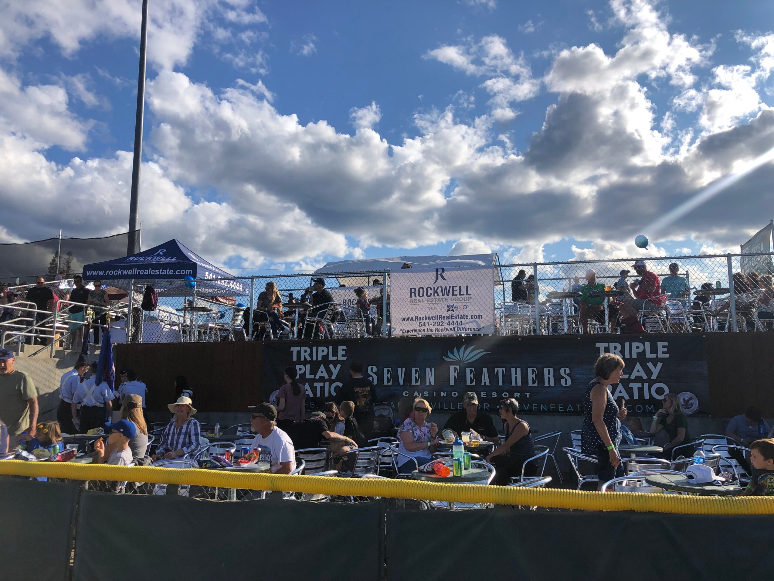 Crowd gathered at an outdoor event with tents, banners, and a stage under a partly cloudy sky.