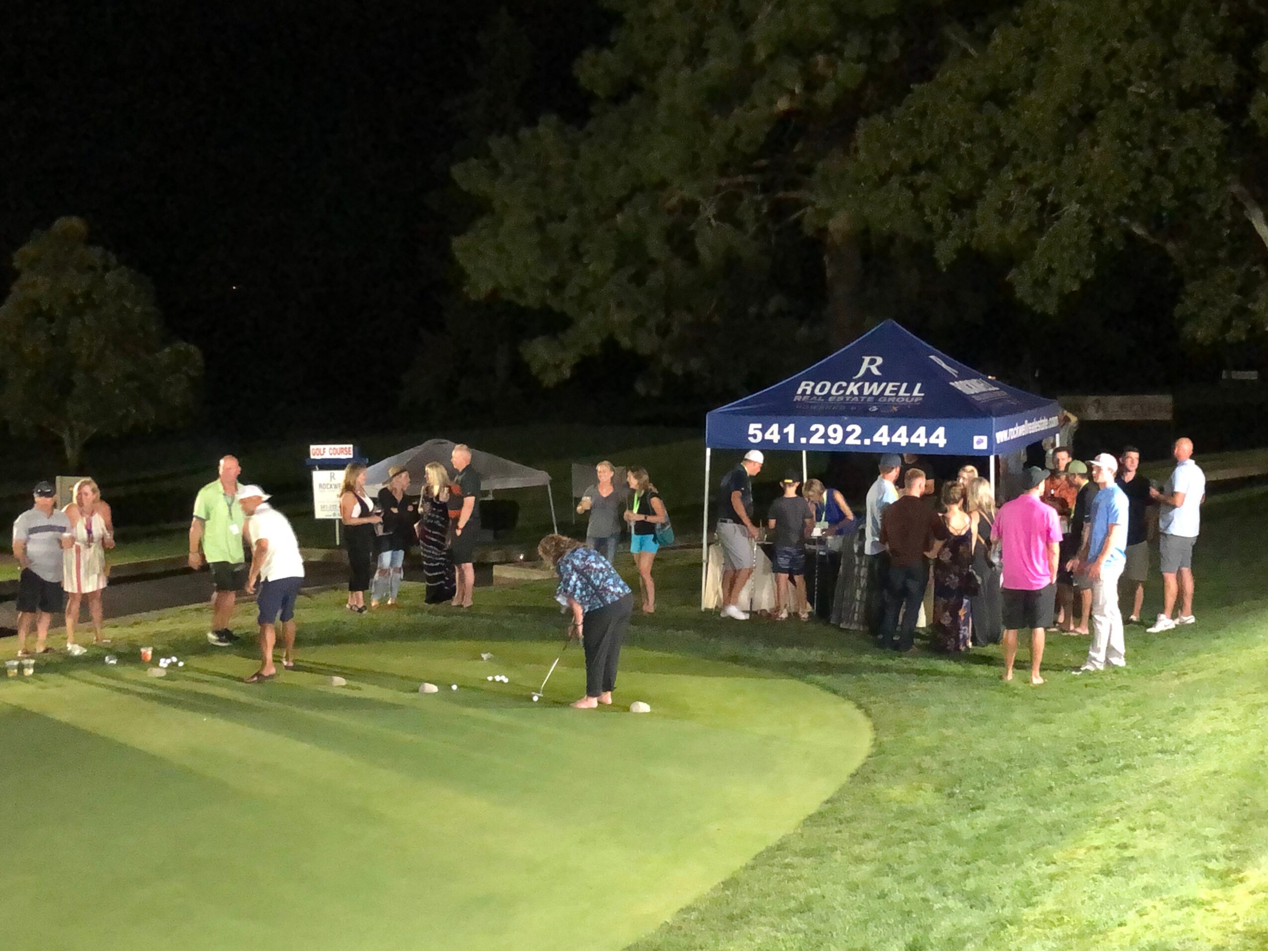 People gather near a tent and trees at night, some playing golf on a green, others socializing under a canopy.