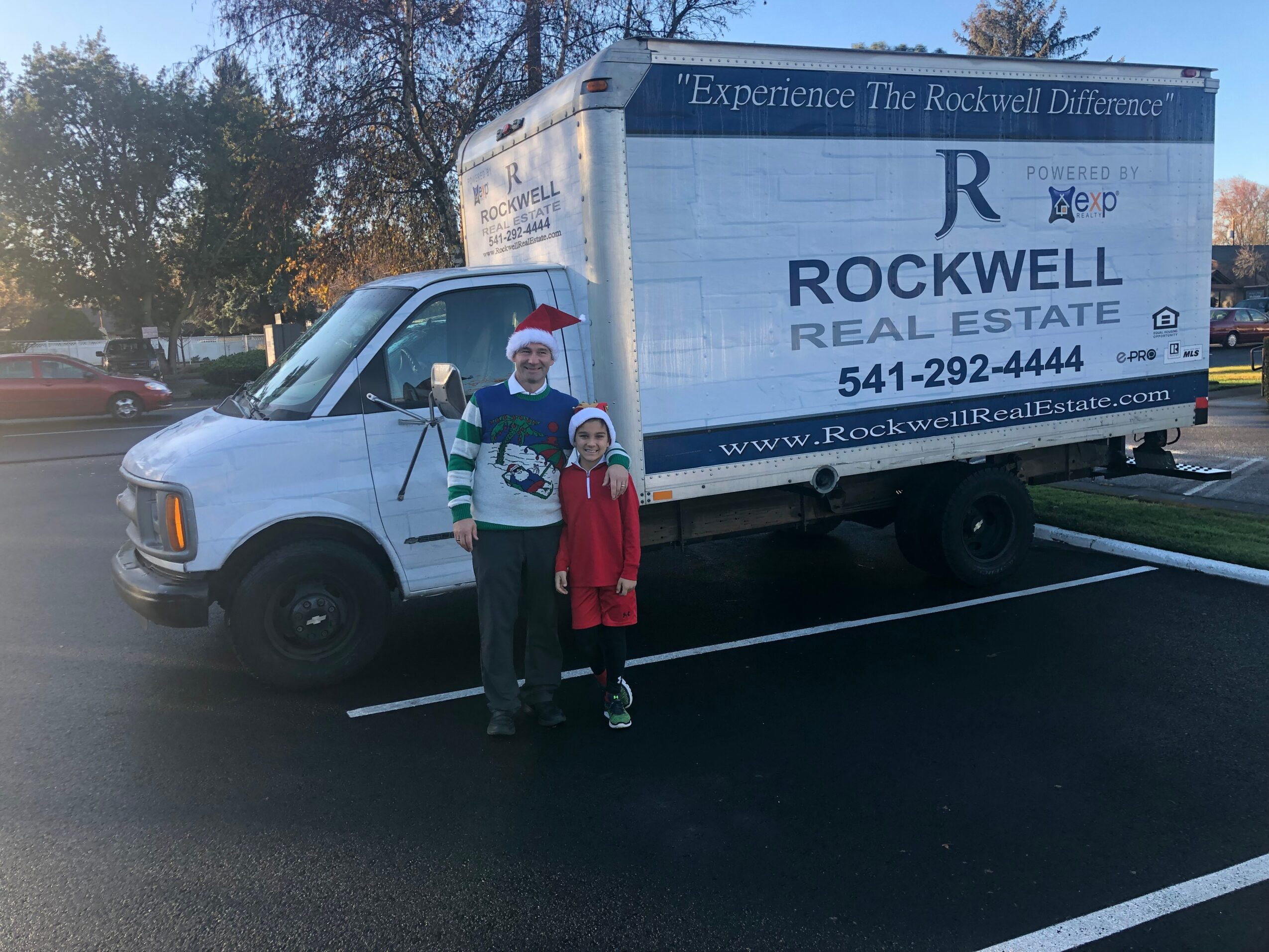 Two people, a man and a child, stand in front of a white moving truck with blue and black text, in a parking lot.