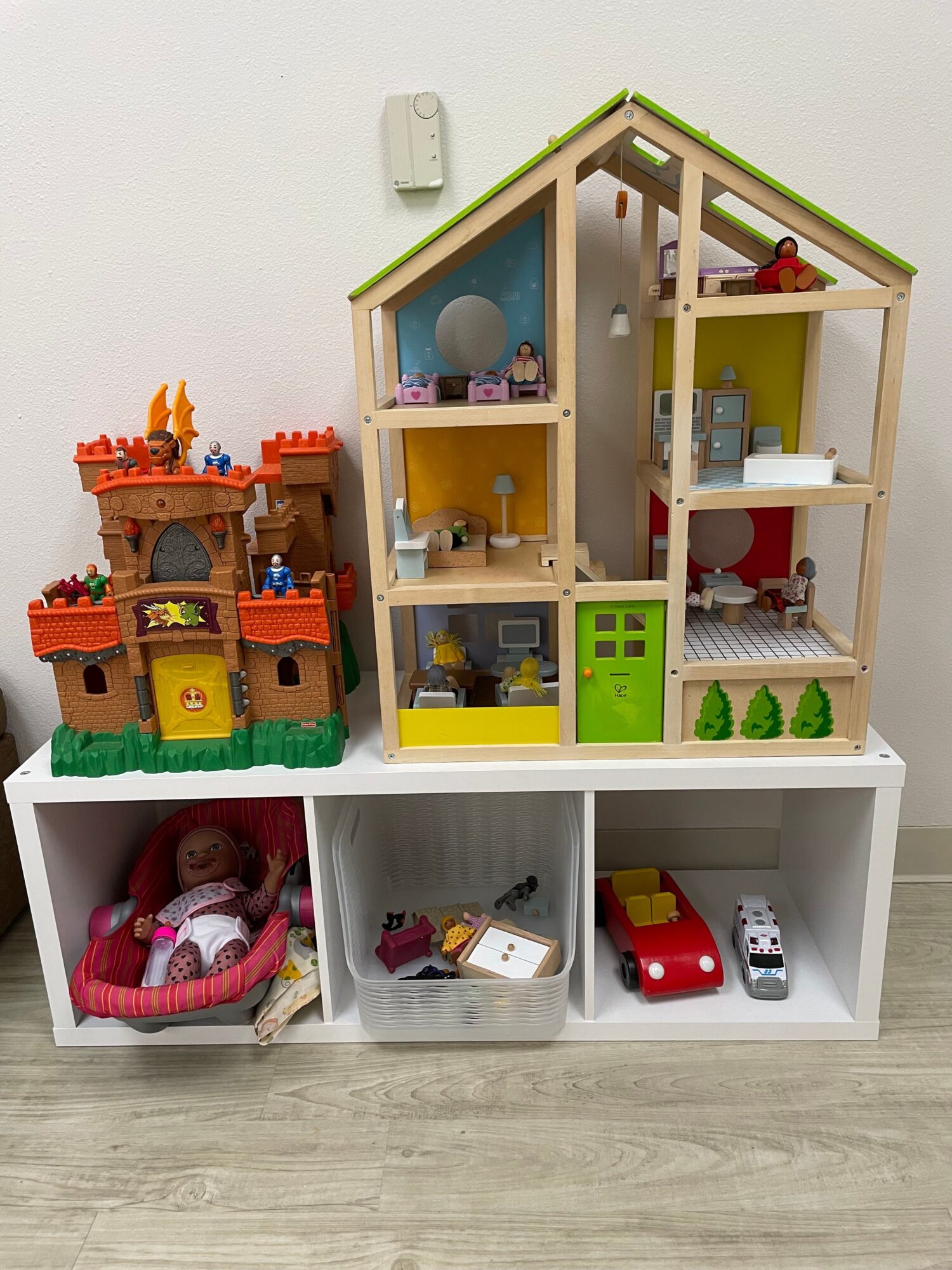 Toy house with multiple rooms, a white shelf with toy vehicles, and a pink doll stroller on a wooden floor.