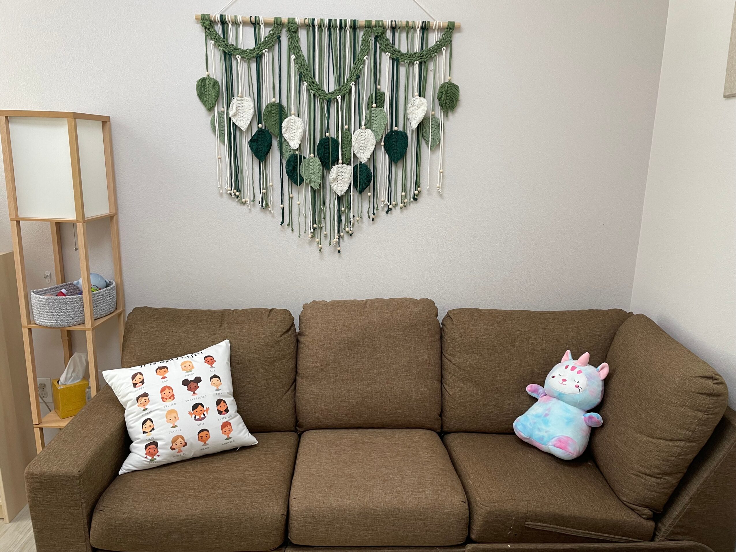 Brown sofa with a decorative pillow and a plush unicorn toy, wall hanging with green and white beads, shelf with glass and basket