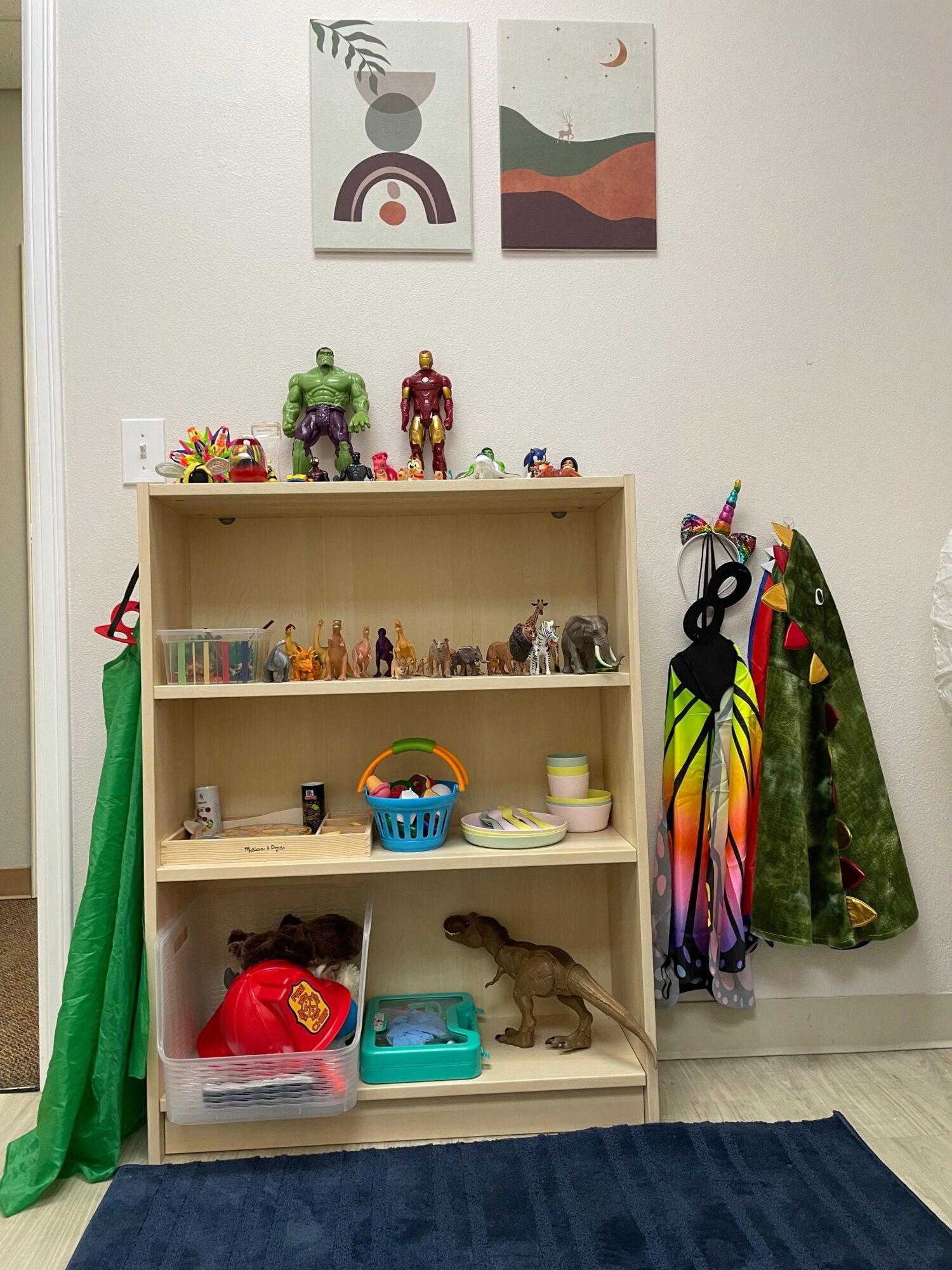 Wooden shelf with toys, books, and decorations, against a white wall with two abstract art pieces, and a green curtain below.