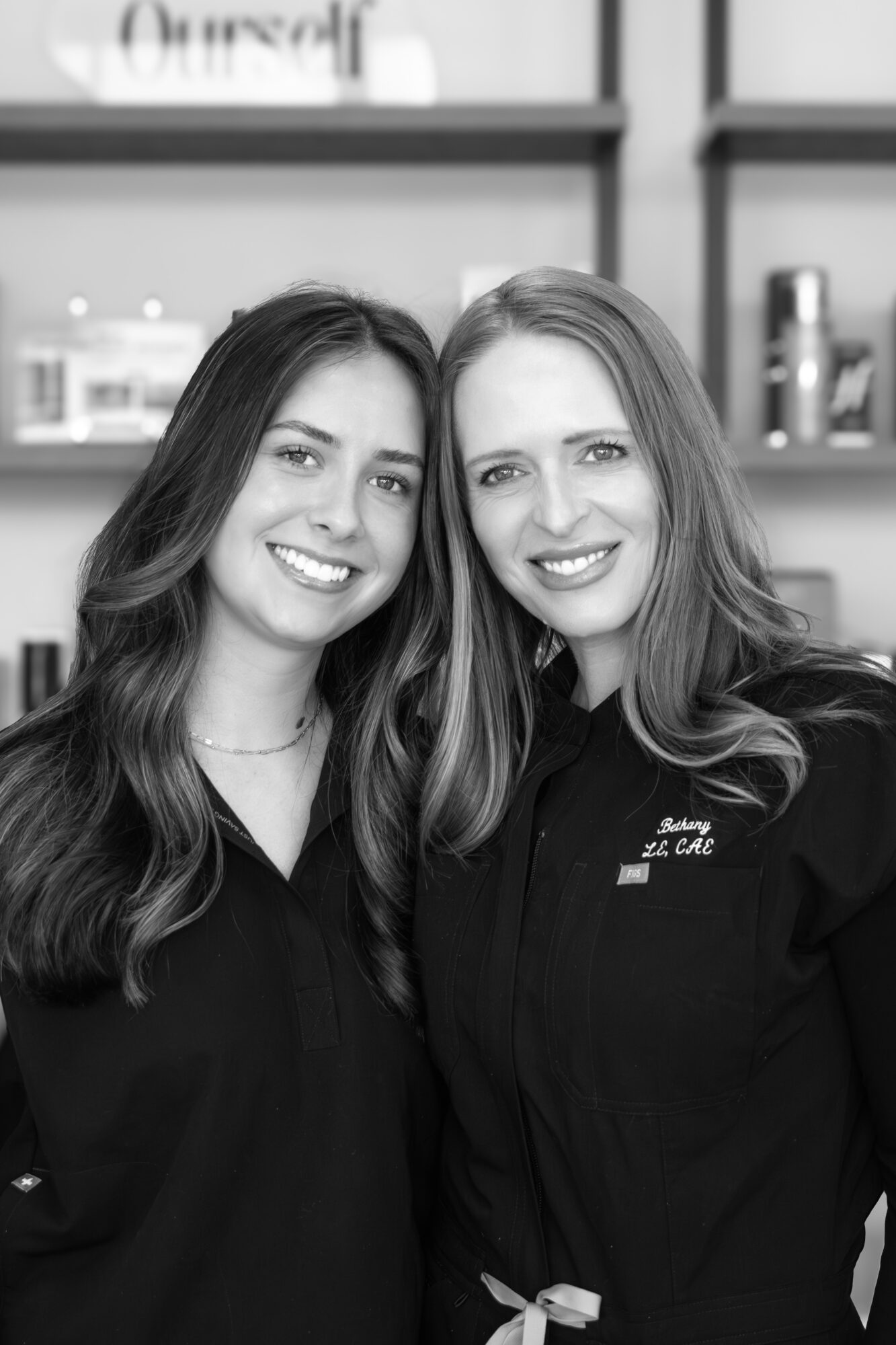 Two women smiling, standing close together indoors, shelves with products in the background.