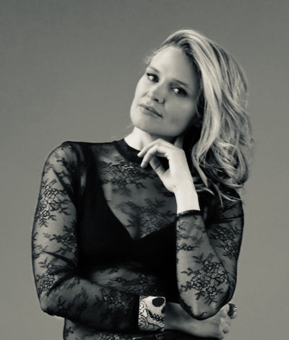 Woman with wavy hair wearing a lace top, resting her chin on her hand, looking at the camera.