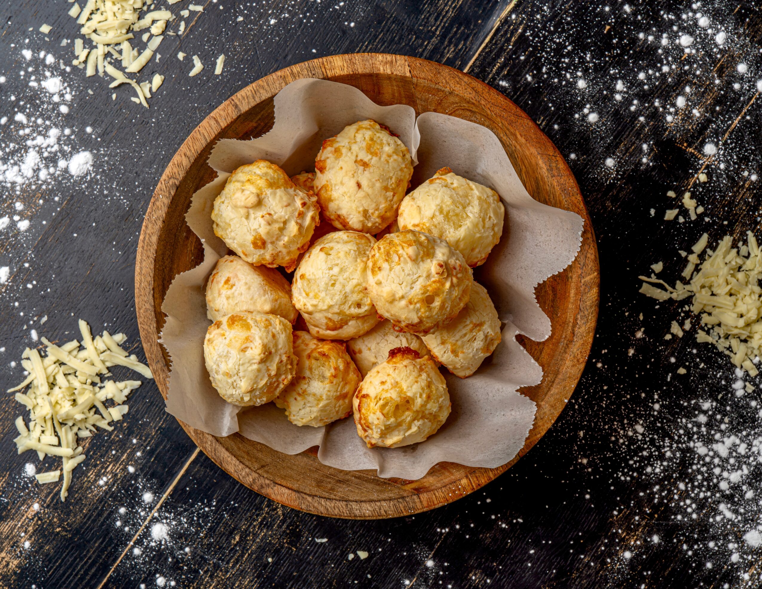 Small round cheese balls in a wooden bowl lined with paper, on a dark surface with scattered cheese shreds and sparkles.