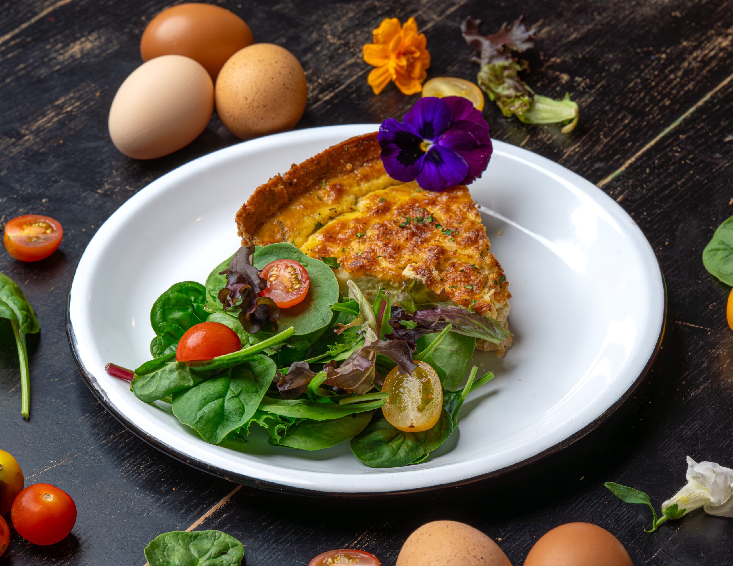 A plate with a slice of quiche and mixed greens, garnished with edible flowers, on a dark wooden surface.
