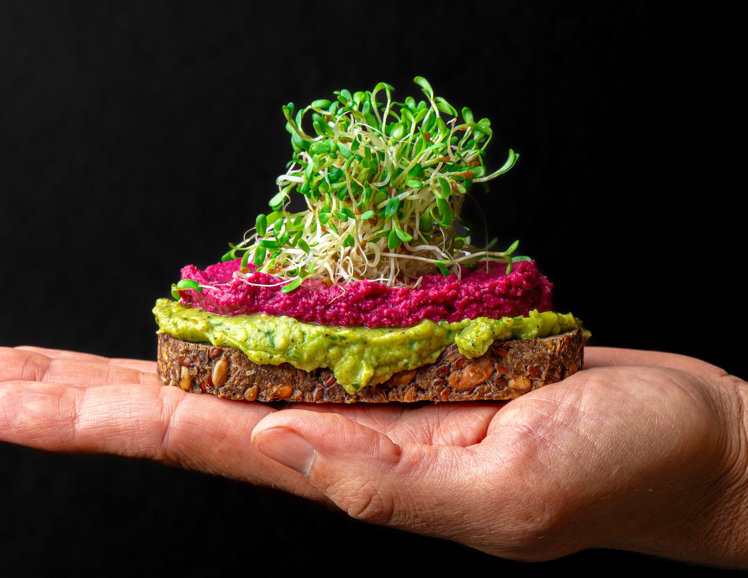 Hand holding a slice of bread topped with green spread, pink topping, and sprouts against a black background.