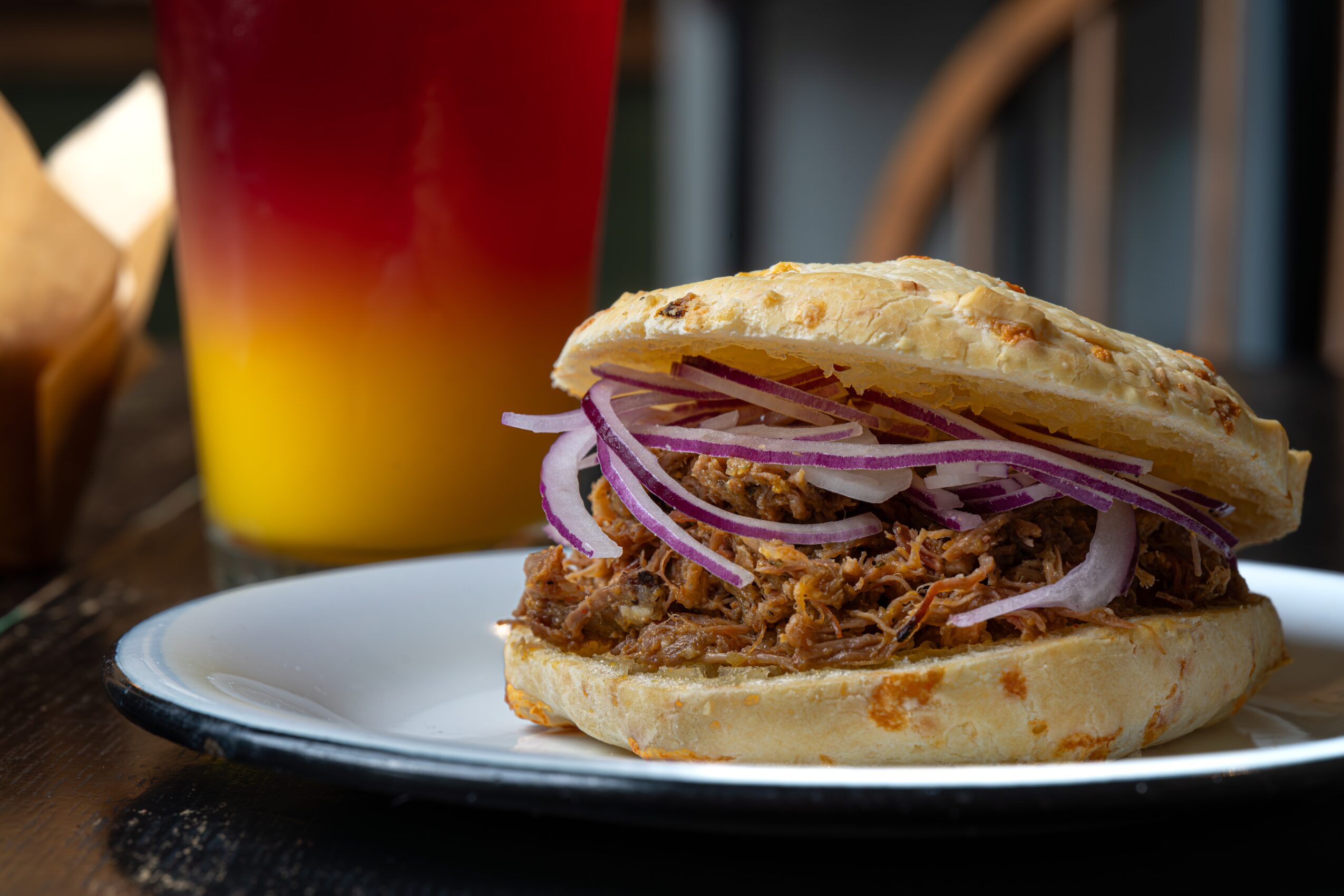 Pulled pork sandwich with onions on a plate, with a colorful drink in the background, in a restaurant setting.