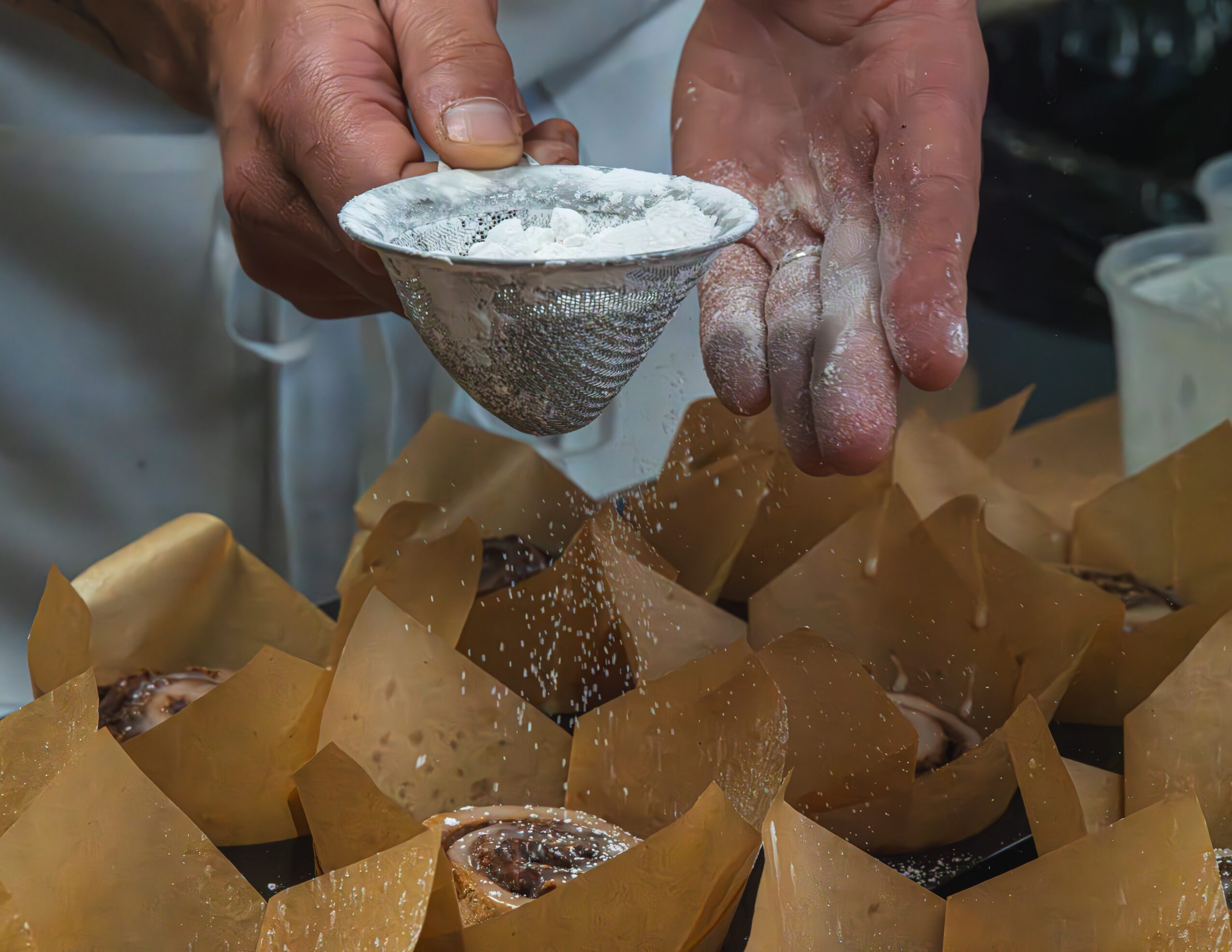 Hands dusting powdered sugar over baked goods wrapped in paper cups, with a sieve in use.