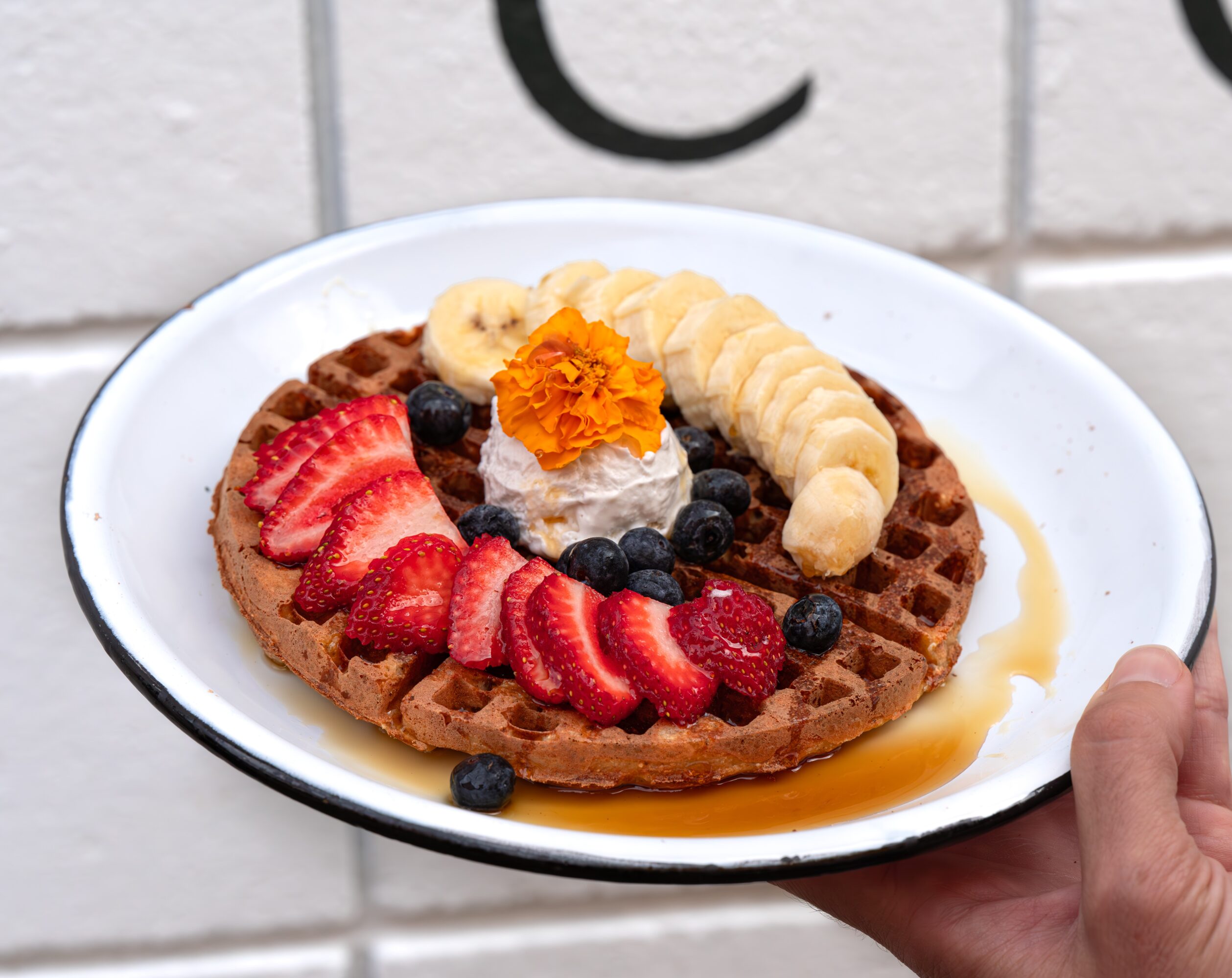 Waffle topped with sliced bananas, strawberries, blueberries, whipped cream, and an orange flower, with syrup on the side, held by a hand.